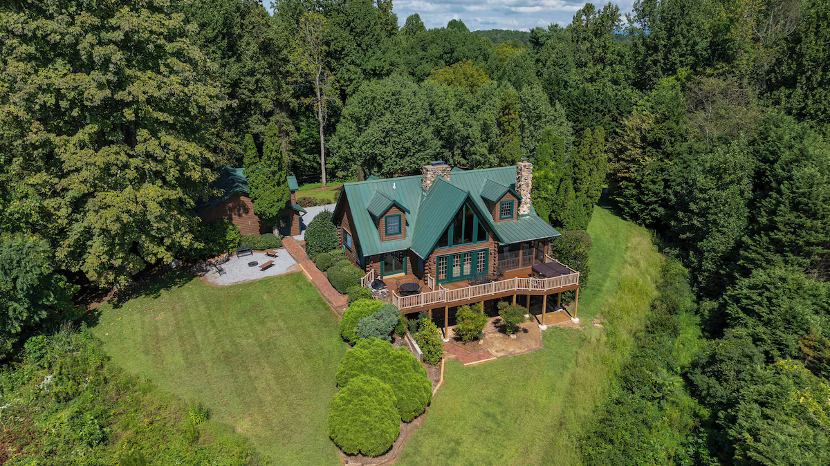 Aerial view of a house with a green metal roof surrounded by trees and a fenced backyard with a patio and lush lawn.