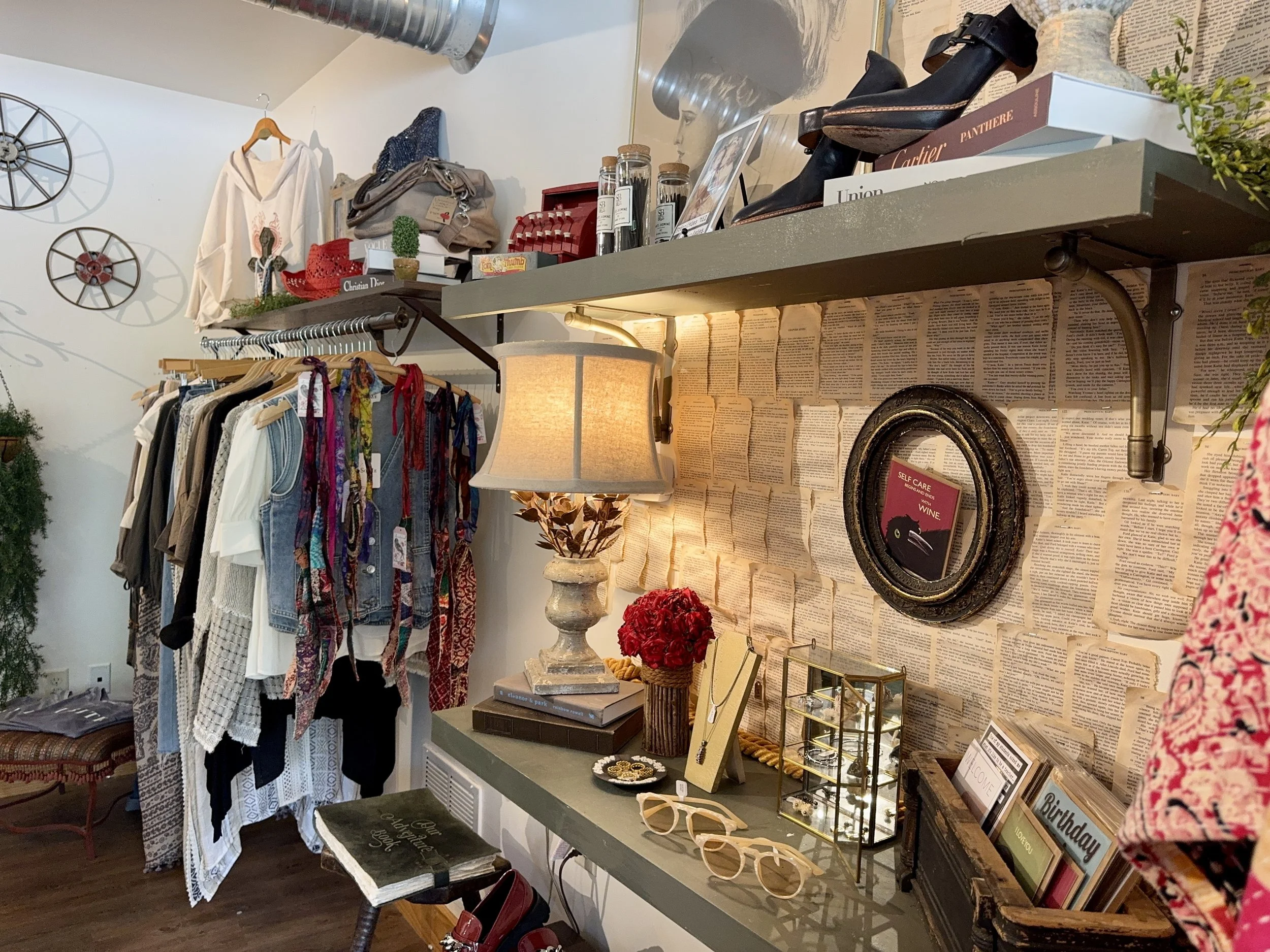 A display of vintage and modern home decor and clothing. On the left, a rack of assorted clothes, including dresses and tops. Above the rack, there are wall decorations with circular metal designs. On the right, shelves hold books, shoes, glass jars,
