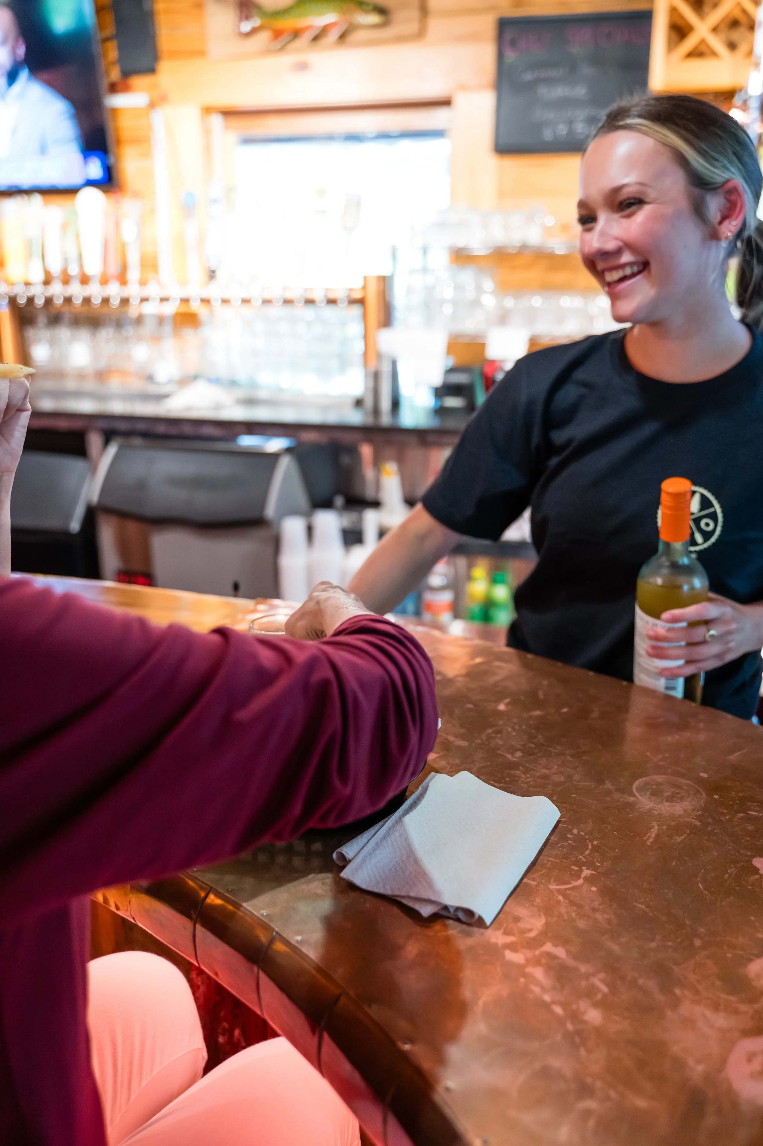 A woman with blonde hair tied in a ponytail, smiling and wearing a black shirt, is holding a bottle of yellow drink at a bar or restaurant counter. In the foreground, a person wearing a maroon long-sleeve shirt is reaching out. The background feature