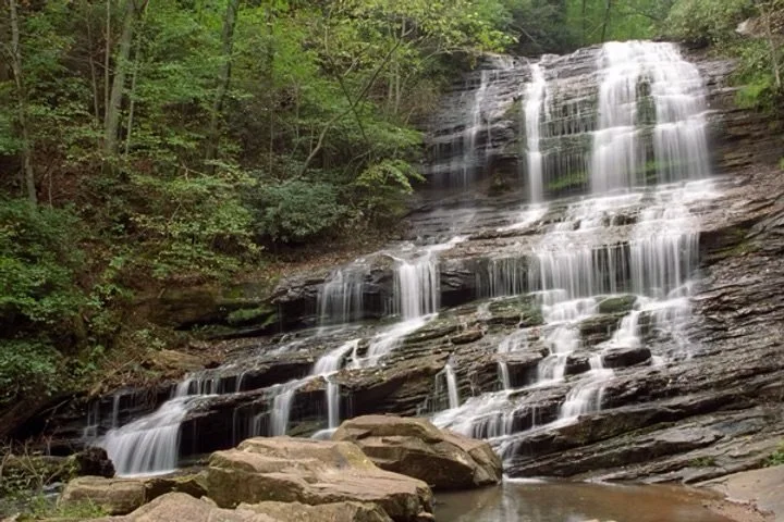 A multi-tiered waterfall flowing over rocks surrounded by green trees in a forest.