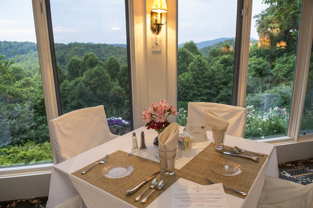 Empty dining table set for four in front of large windows overlooking a lush green landscape with trees and distant hills, with pink flowers in a small vase and a wall-mounted light above.