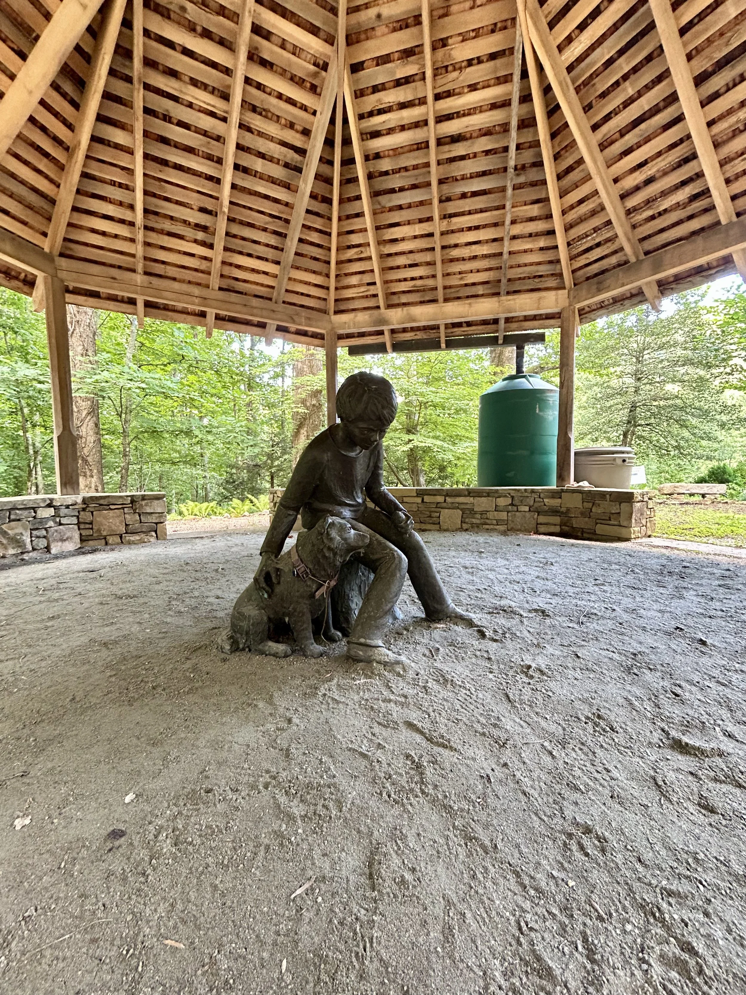 A bronze statue of a boy petting a dog inside a wooden pavilion in a park, with trees and greenery outside.