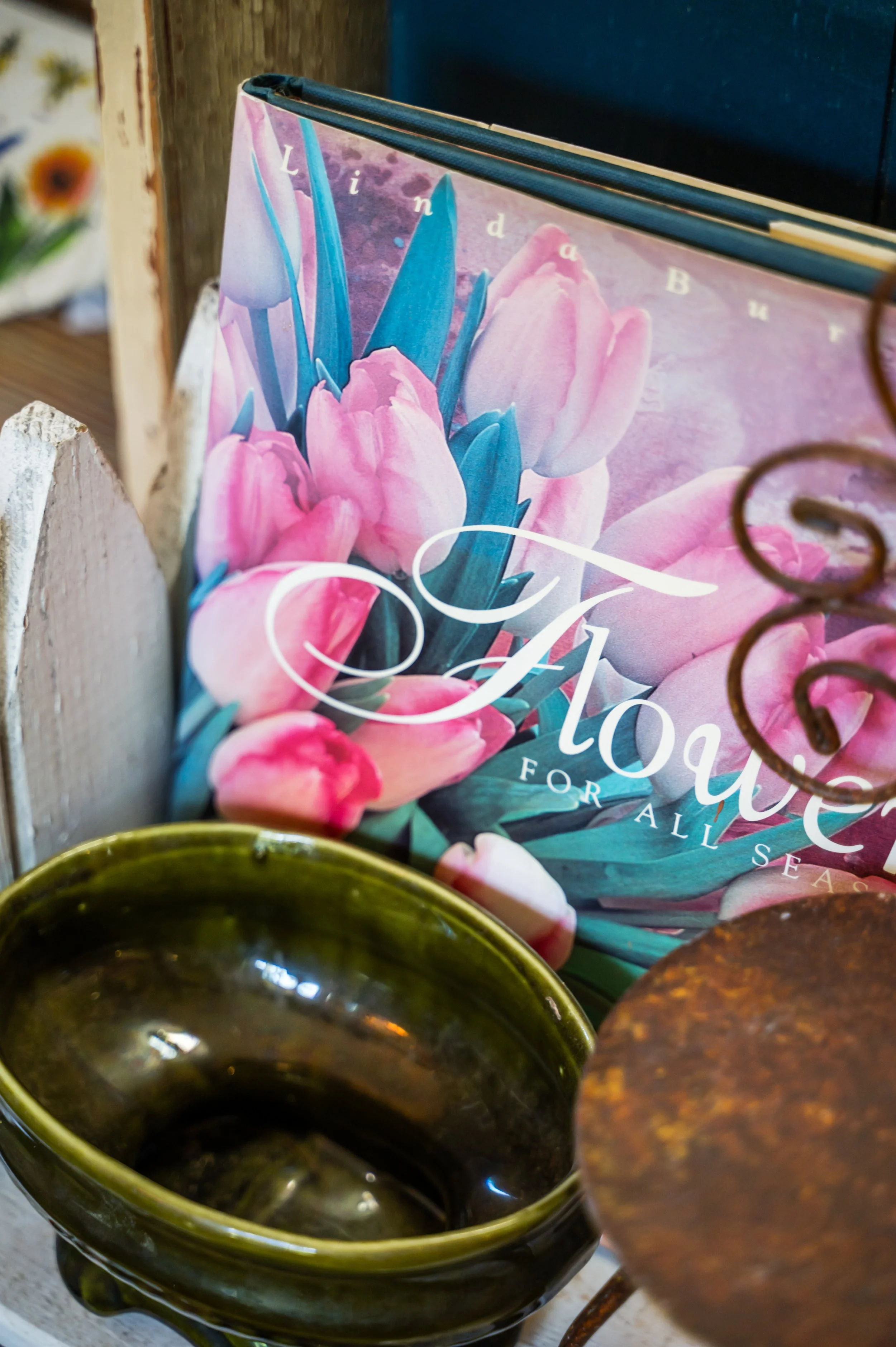 Close-up of a dark green ceramic cup filled with a dark liquid, likely coffee, placed on a weathered wooden surface. Partially visible is a floral book or magazine with pink and blue flowers on the cover behind the cup.