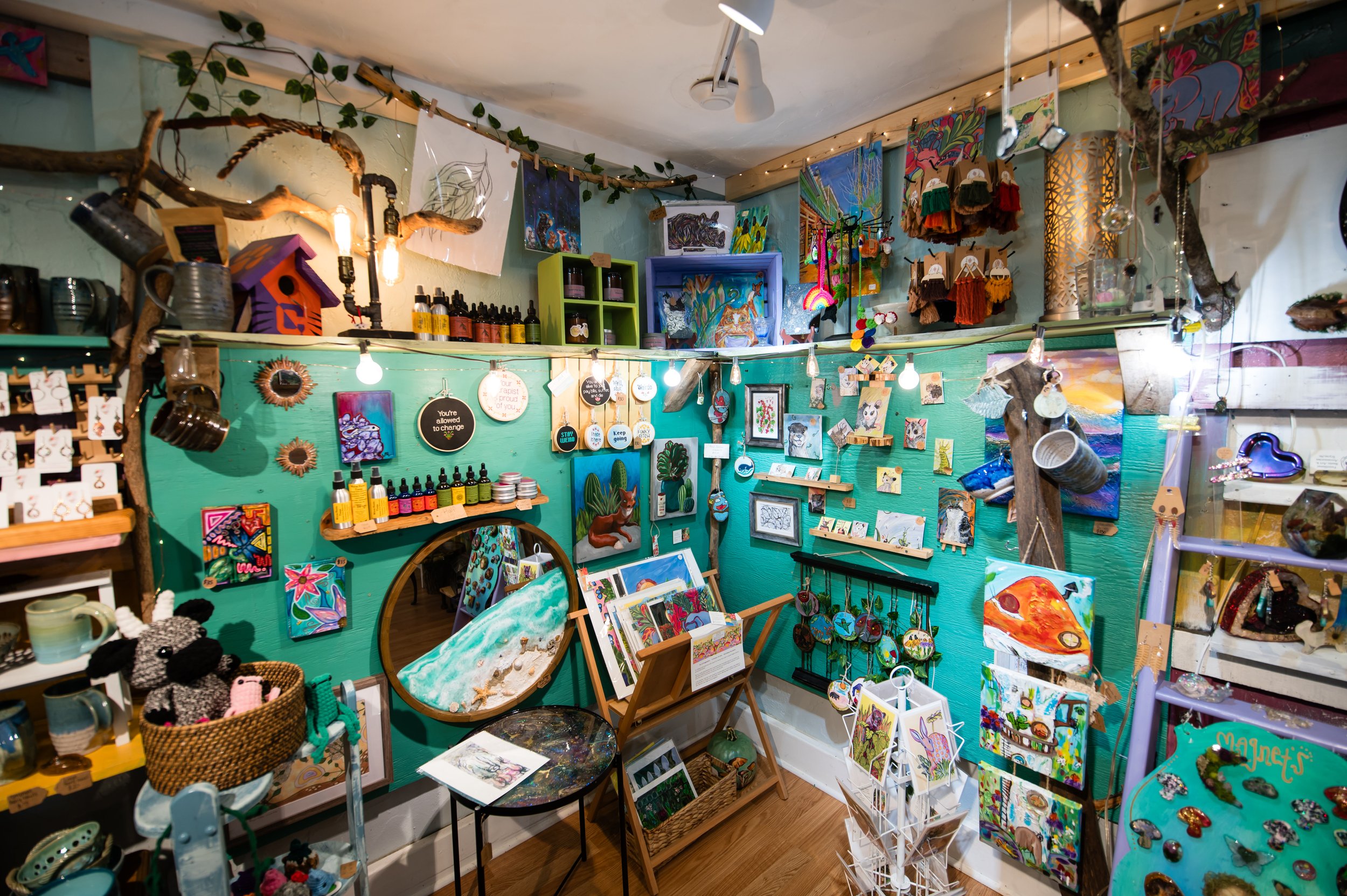 A wall display of colorful artwork, paintings, and handmade crafts at an art or craft store, with shelves, hanging decorations, and various art supplies visible.
