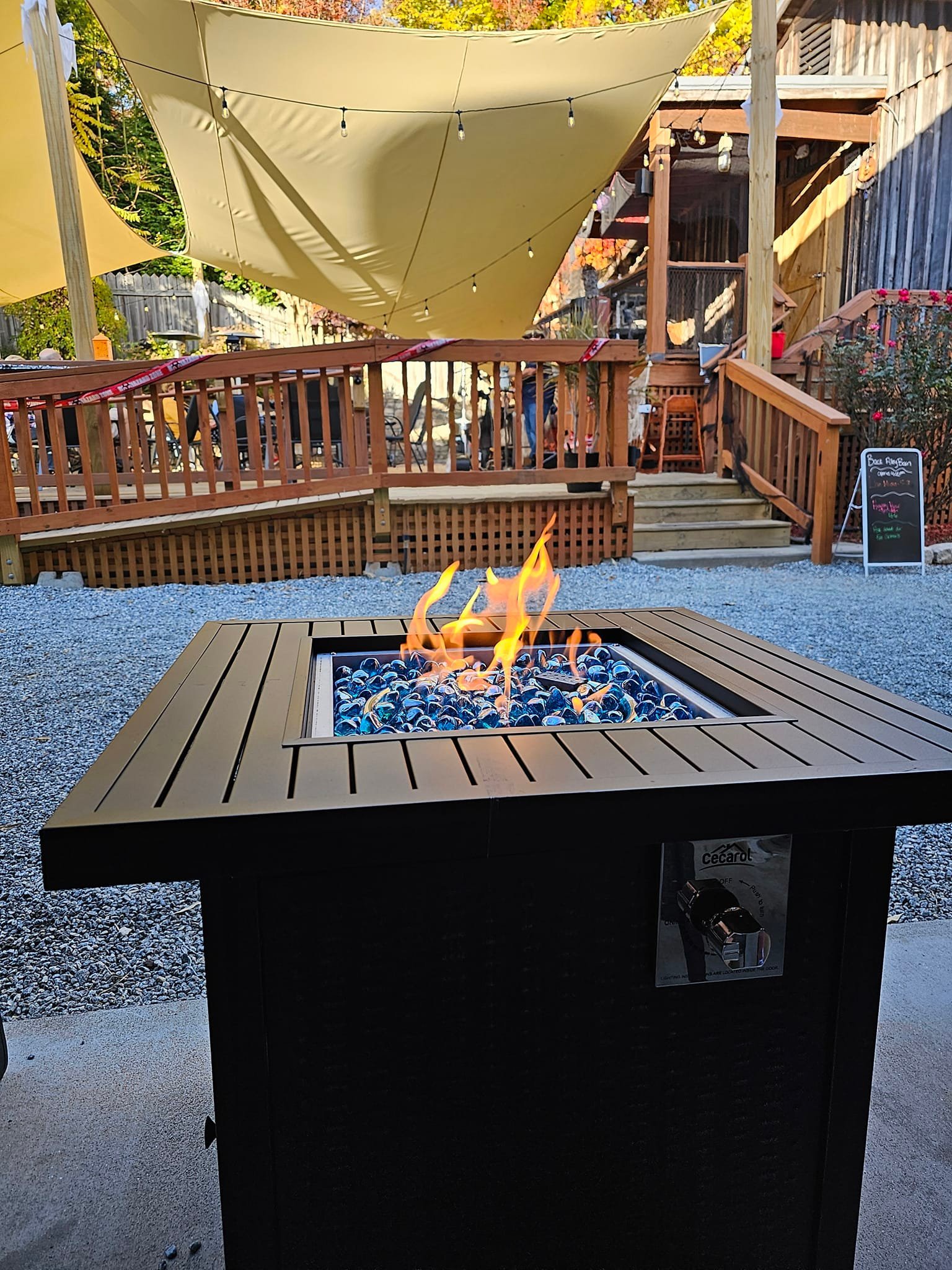 Outdoor patio with a fire pit in the foreground, a wooden deck with a yellow canopy and string lights above, surrounded by trees with fall foliage, and a chalkboard sign on the right side.