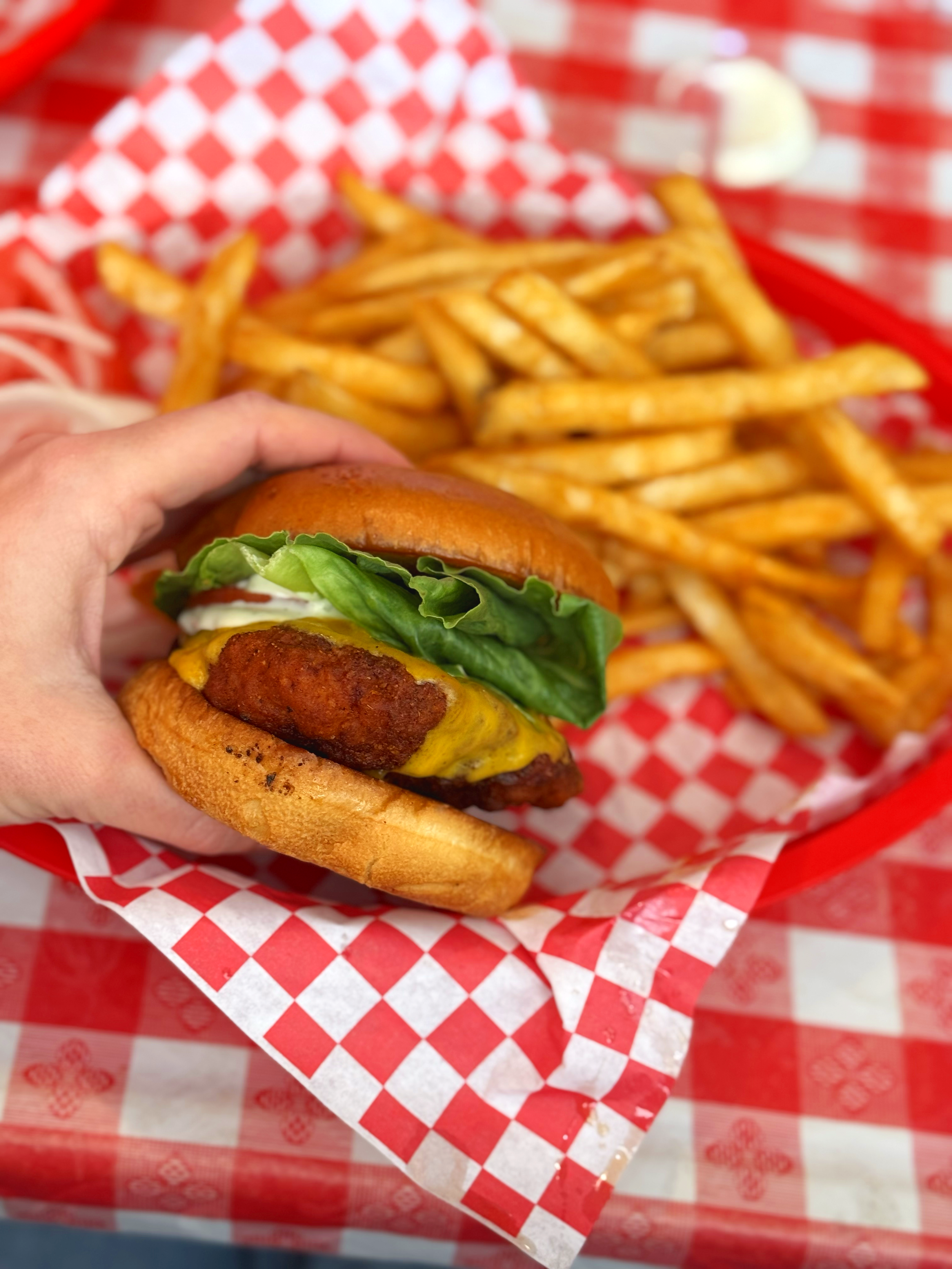 A cheeseburger with lettuce, tomato, and a fried chicken patty, along with a side of French fries in a red basket with checkered paper.