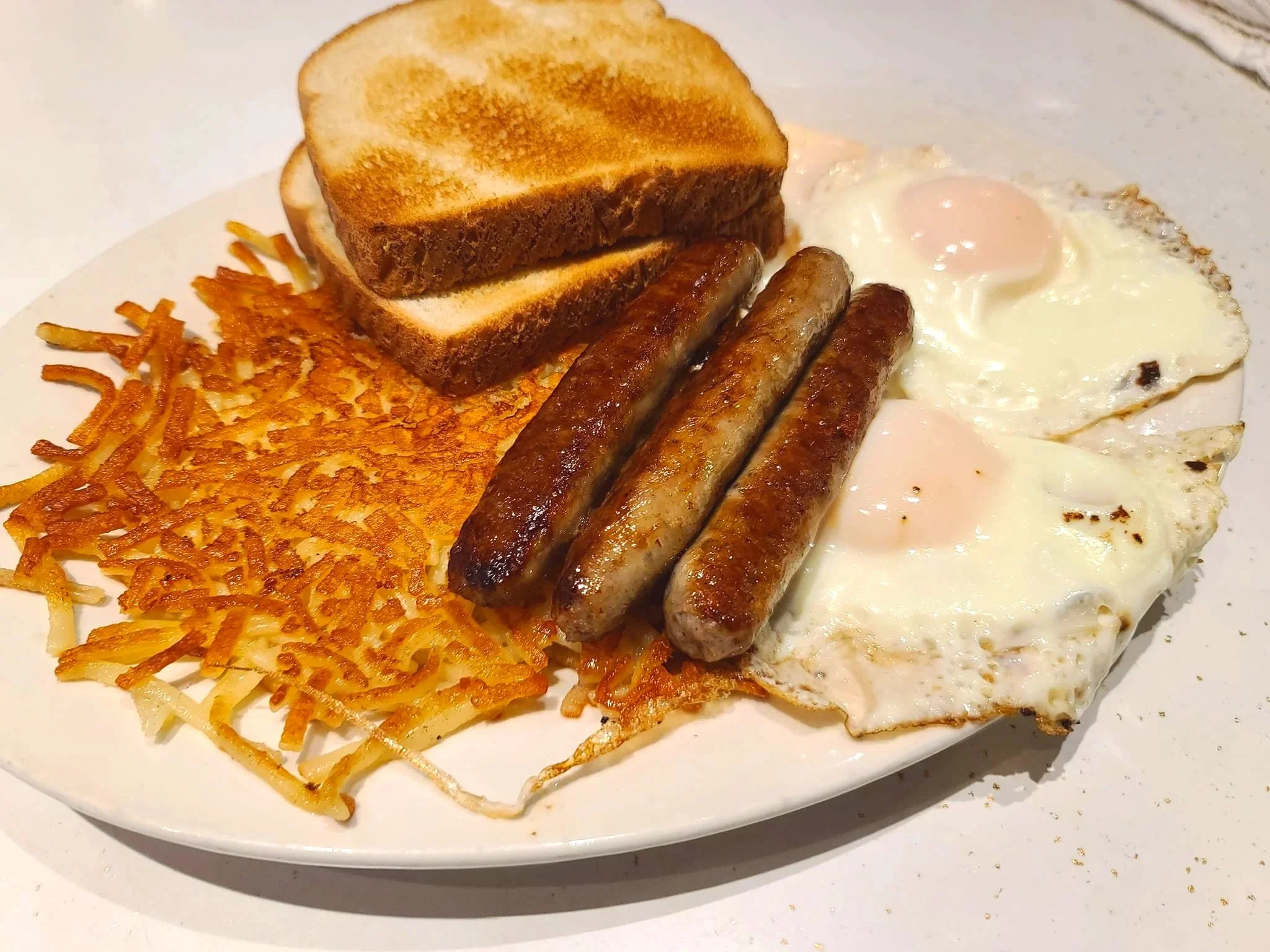 A white plate with fried hash browns, three cooked sausages, two fried eggs, and toasted bread slices.