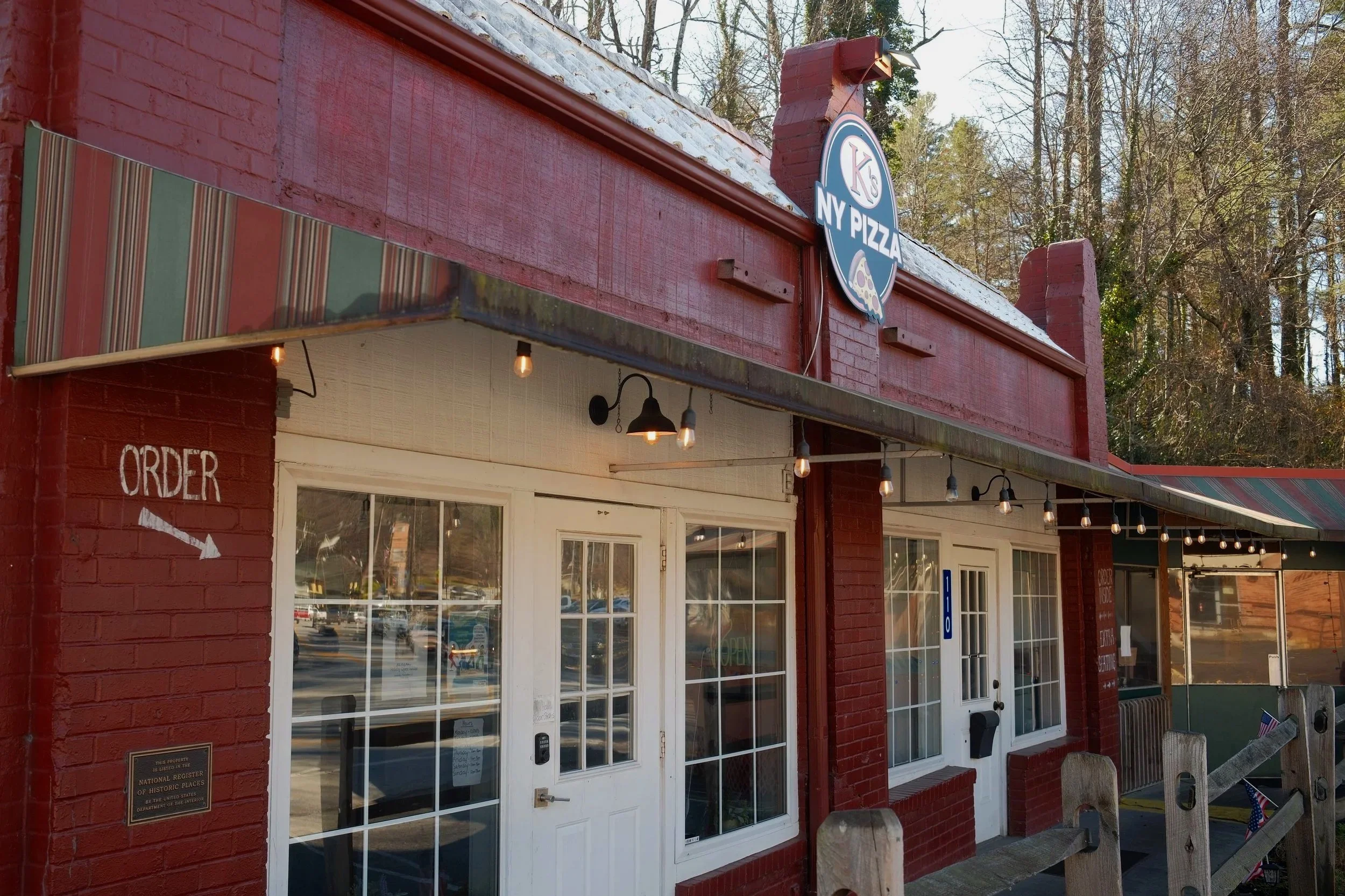 The facade of a red brick building with a sign that reads "NY PIZZA" and a pizza graphic. Below the sign, there is an entrance with glass doors and windows, some string lights hanging, and a small fence with patriotic decorations.