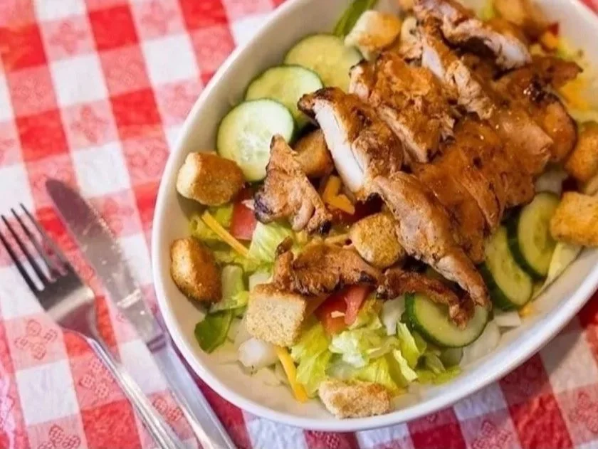 A bowl of chicken Caesar salad with grilled chicken, croutons, cucumbers, shredded cheese, and chopped lettuce, on a red and white checkered tablecloth.