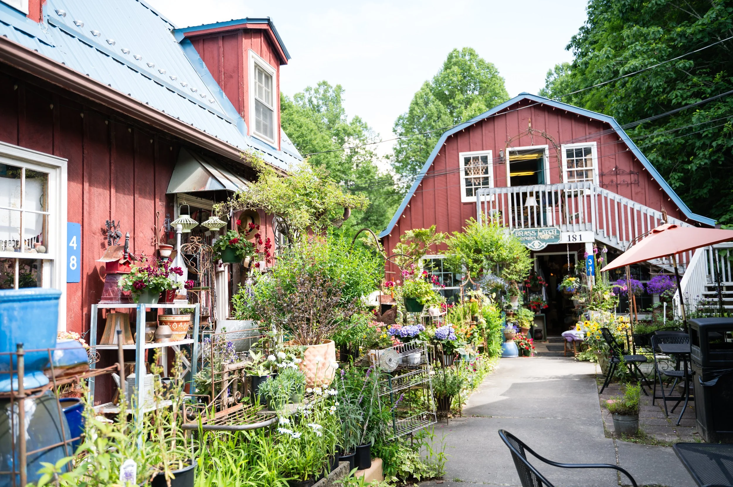 Colorful garden shop with various plants and flowers outside, red wooden buildings with blue metal roofs, outdoor seating, and green trees in the background.