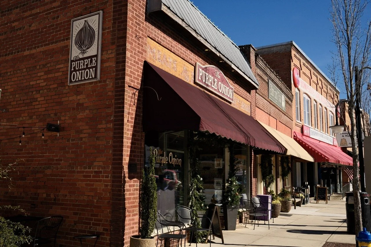 Street view of storefronts with red brick buildings and various awnings, including 'The Purple Onion,' decorated for the holidays with small trees and plants outside.