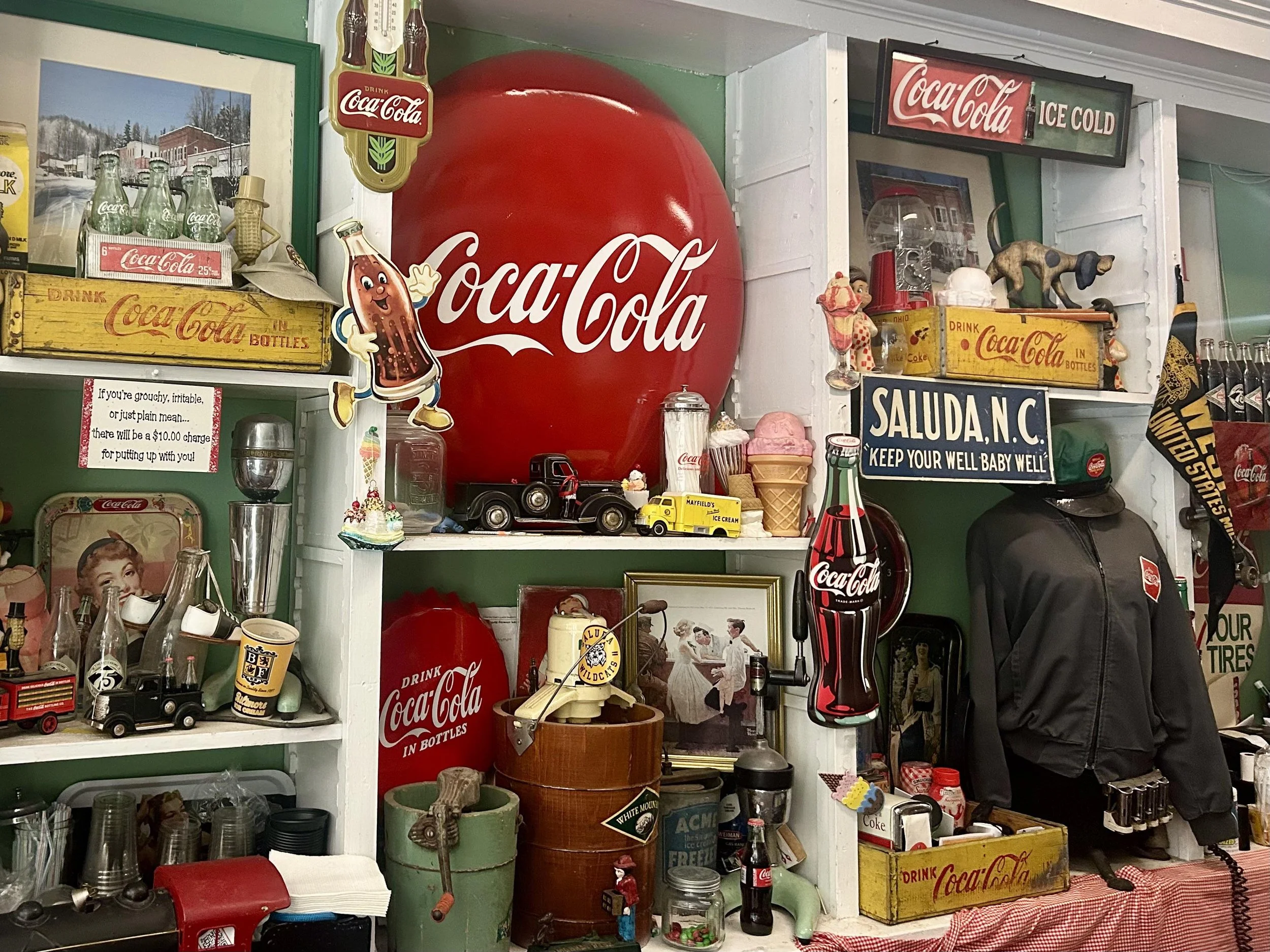 A collection of vintage Coca-Cola signs, bottles, and memorabilia on white shelves, including a large red Coca-Cola balloon, toy trucks, ice cream cone decor, and various retro advertising items.
