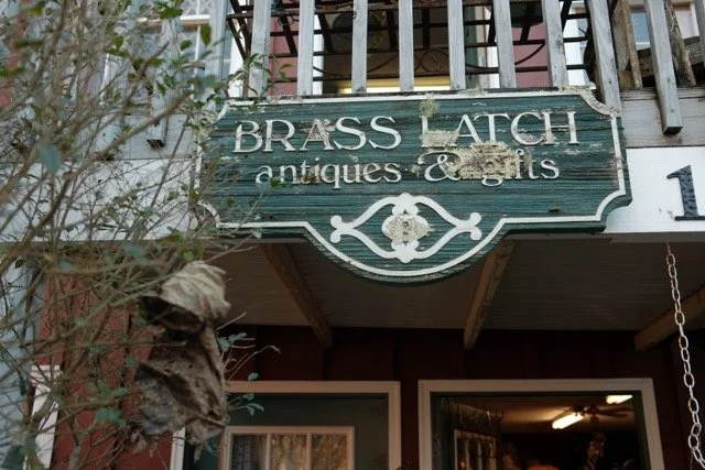 Green weathered wooden sign reading 'Brass Latch antiques & gifts' hanging above a store entrance.