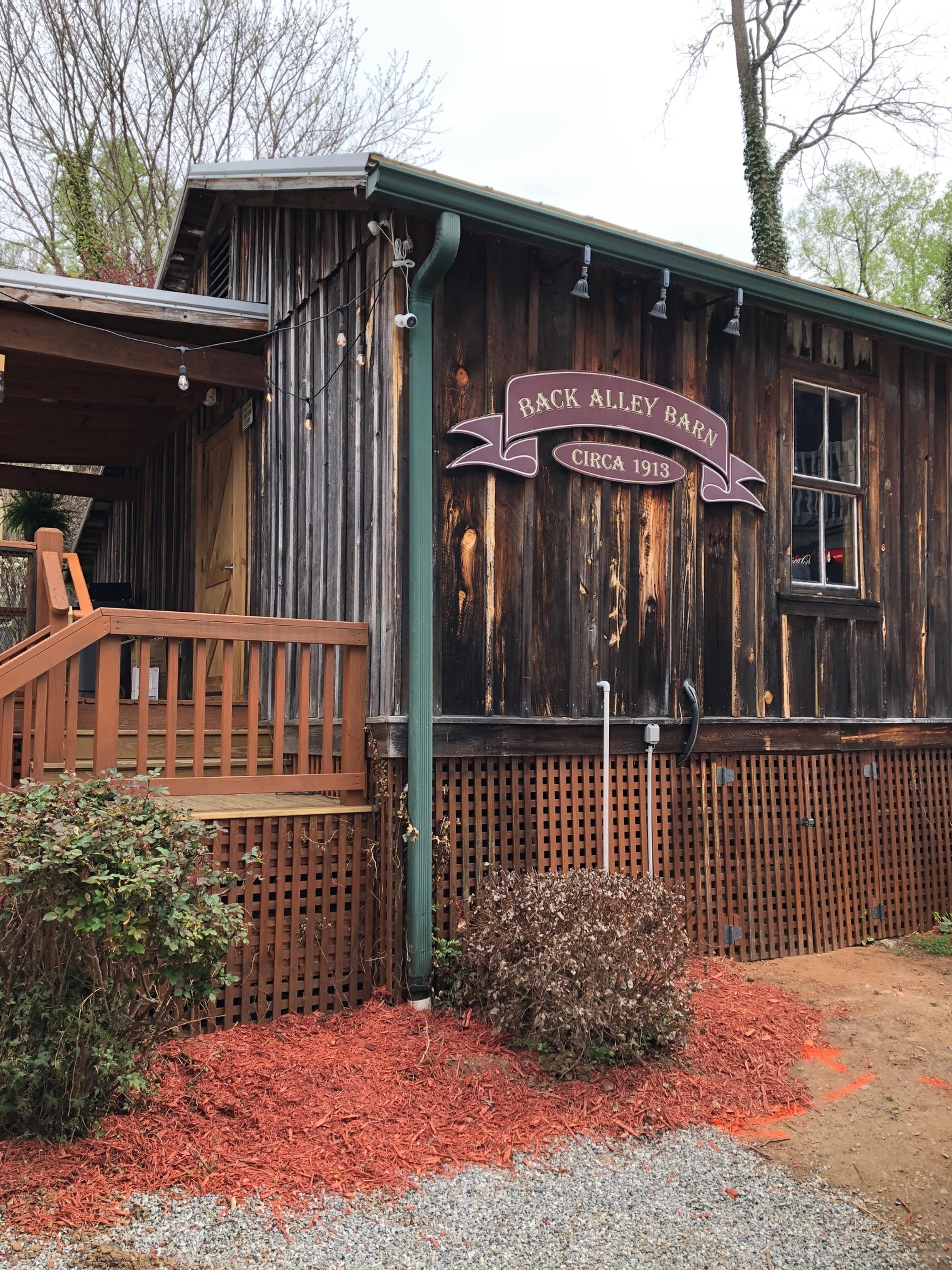 A rustic wooden barn with a sign that reads 'Back Alley Barn Circa 1913', surrounded by a wooden lattice fence, outdoor string lights, a few bushes, and a gravel pathway with orange wood mulch.