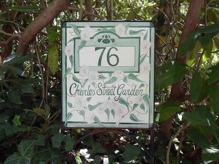House address sign with the number 76 and the text 'Charles Street Garden' surrounded by green ivy and white flowers, mounted on a plant-filled green hedge.