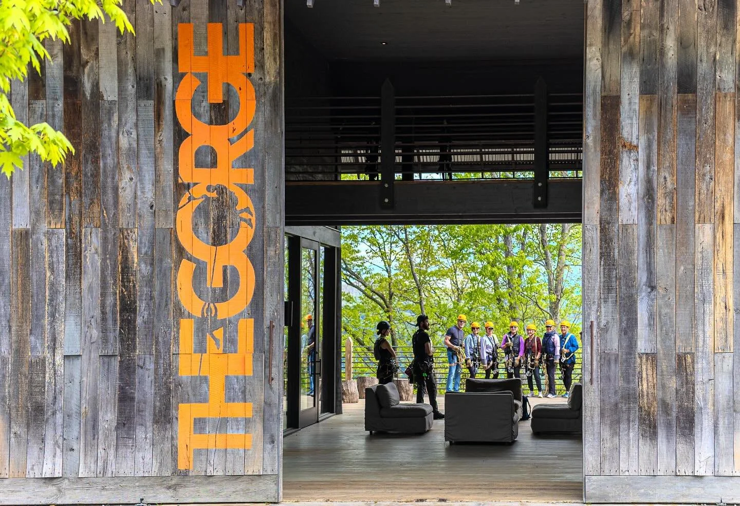 A group of people wearing helmets and safety gear standing outside a building with a rustic wooden exterior at a treetop adventure park.