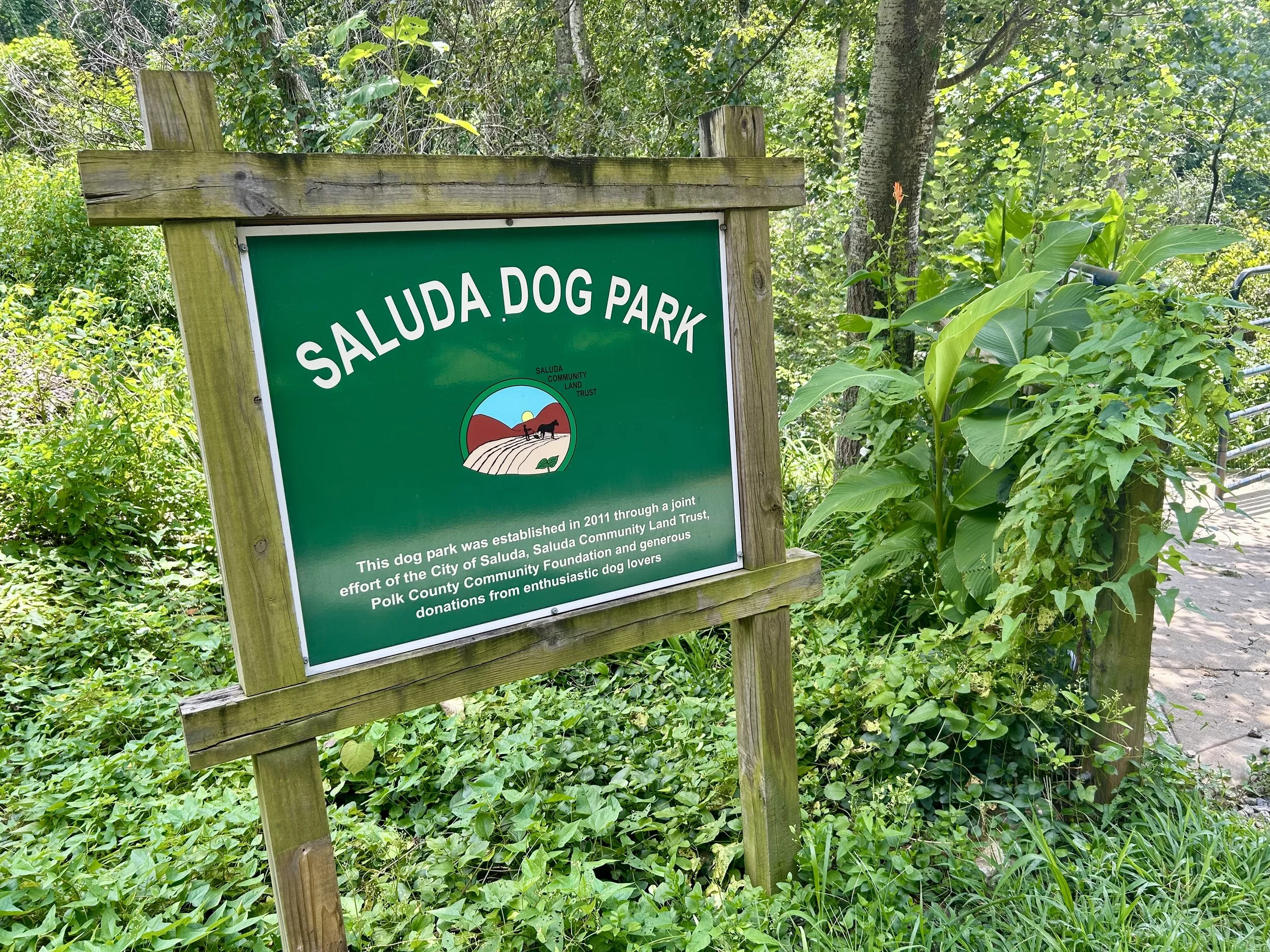 Sign for Saluda Dog Park surrounded by lush green trees and plants.