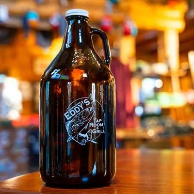 A brown glass growler labeled 'Eddy's Tap Room Grill' on a wooden table in a restaurant or bar setting.