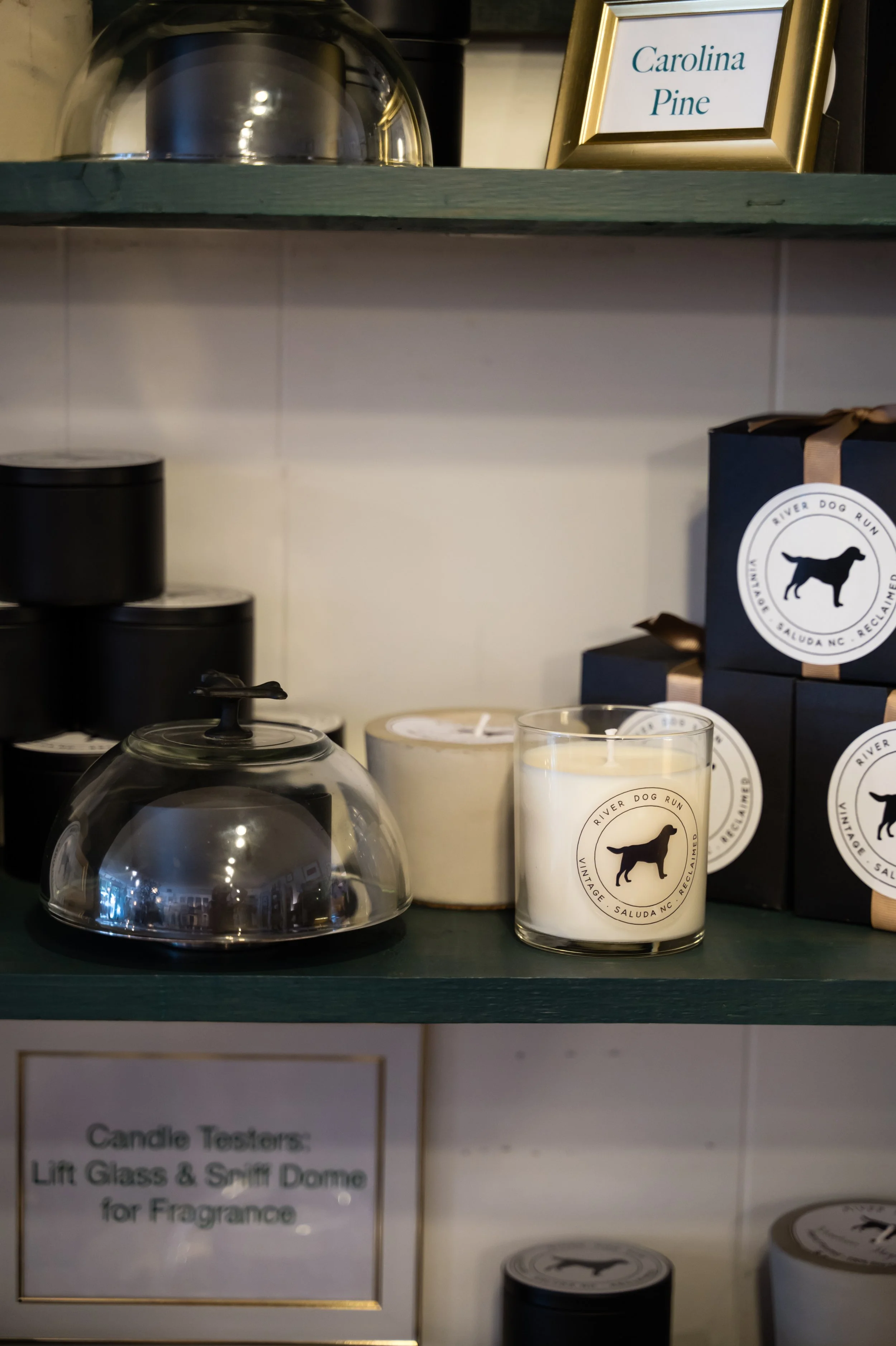 Display shelf with candles and candle testing kit, featuring a framed sign that reads 'Carolina Pine,' and a box with a dog silhouette logo from River Dog Run.