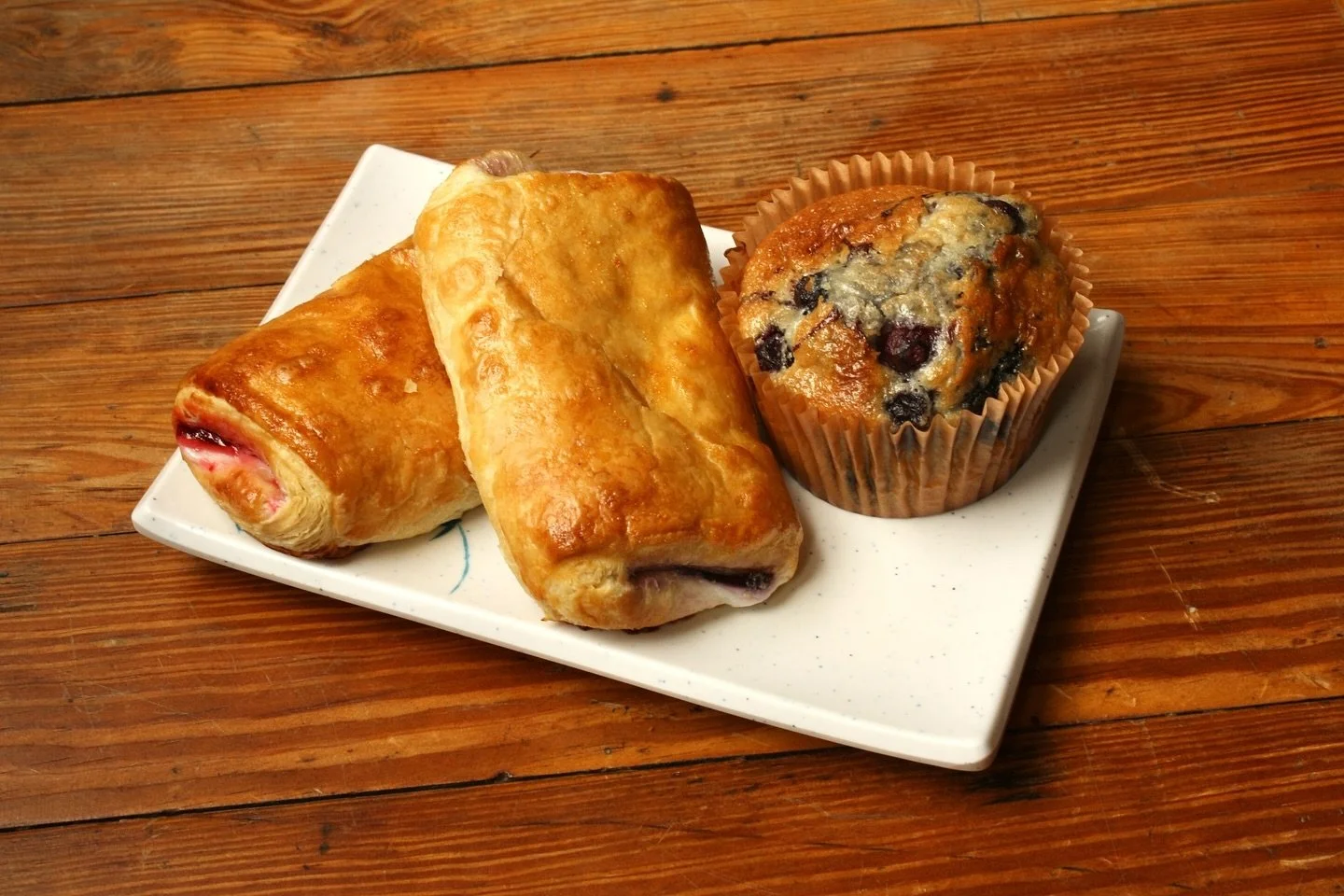 Plate with two ham and cheese croissants and a blueberry muffin on a wooden table.