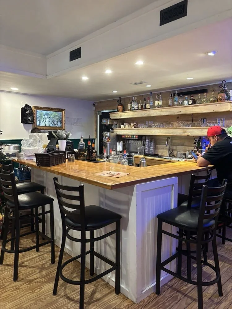 A cozy bar with a wooden counter and black chairs, bottles of alcohol on shelves, a bartender station, and a man sitting at the bar using his phone.