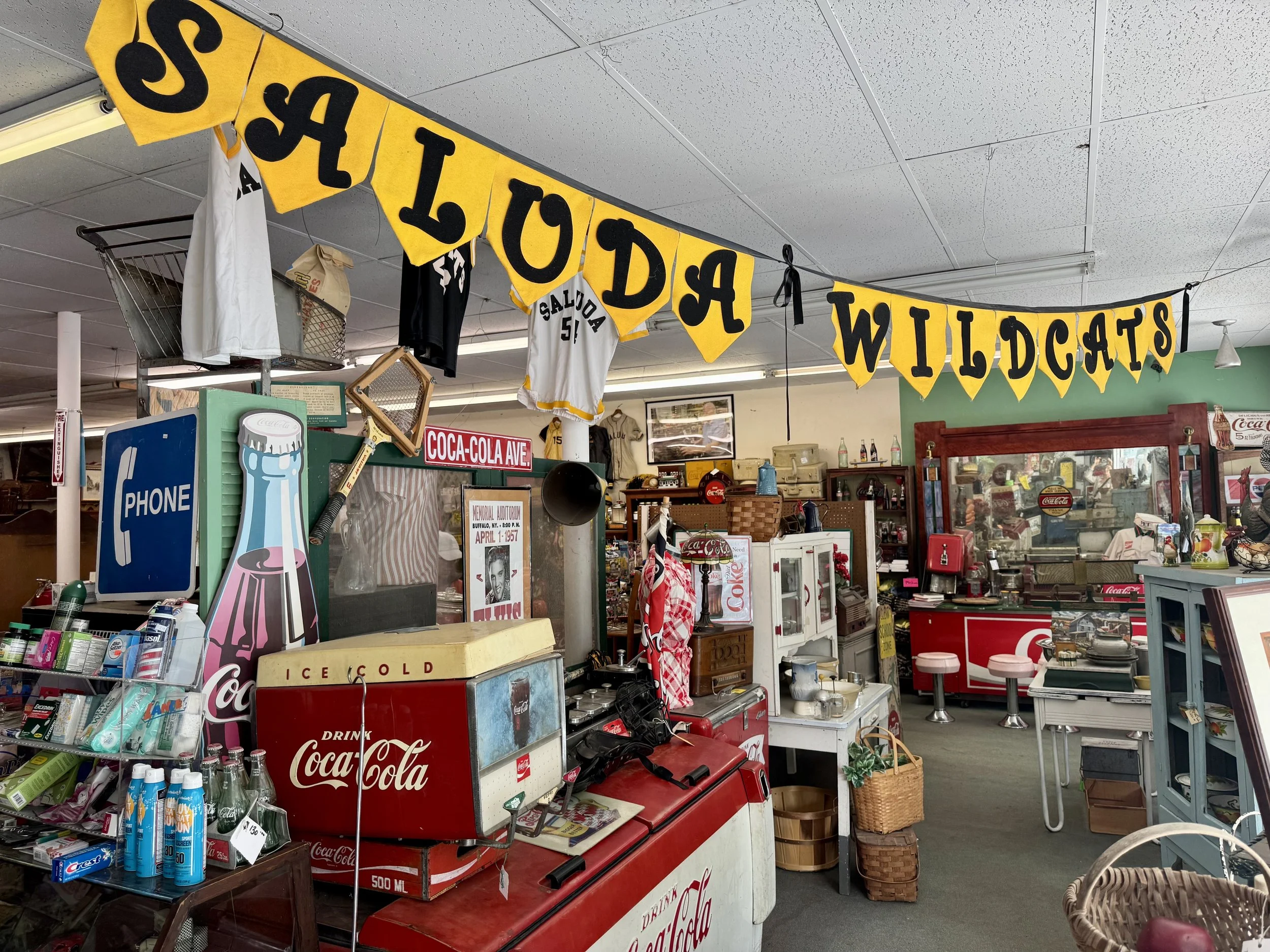 Inside a vintage store with a yellow and black 'GASOLINE WILDCATS' banner hanging from the ceiling. Various antiques, vintage signs, and household items are on display, including soda coolers, baskets, and framed pictures.