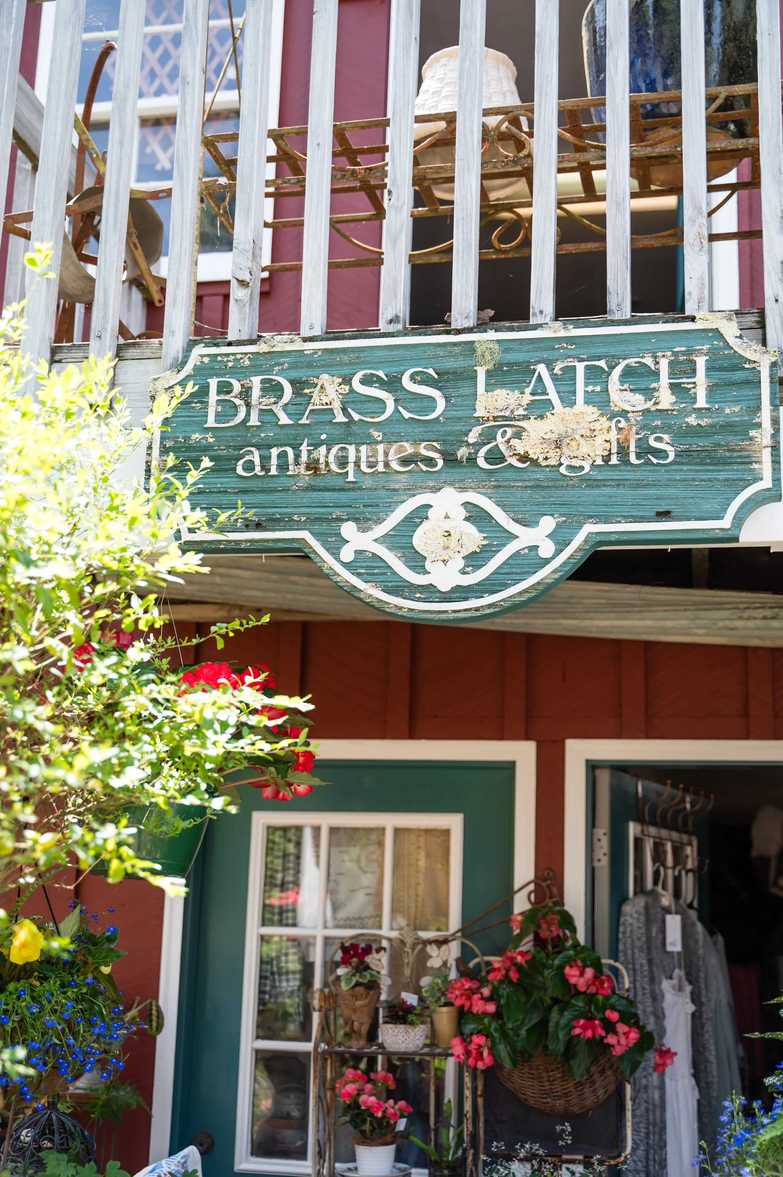 A storefront with a weathered wooden sign that reads 'Brass Laptop antiques & gifts' hangs above a display of colorful potted flowers outside. The storefront has a teal door and window, with various decorative items and plants visible inside.