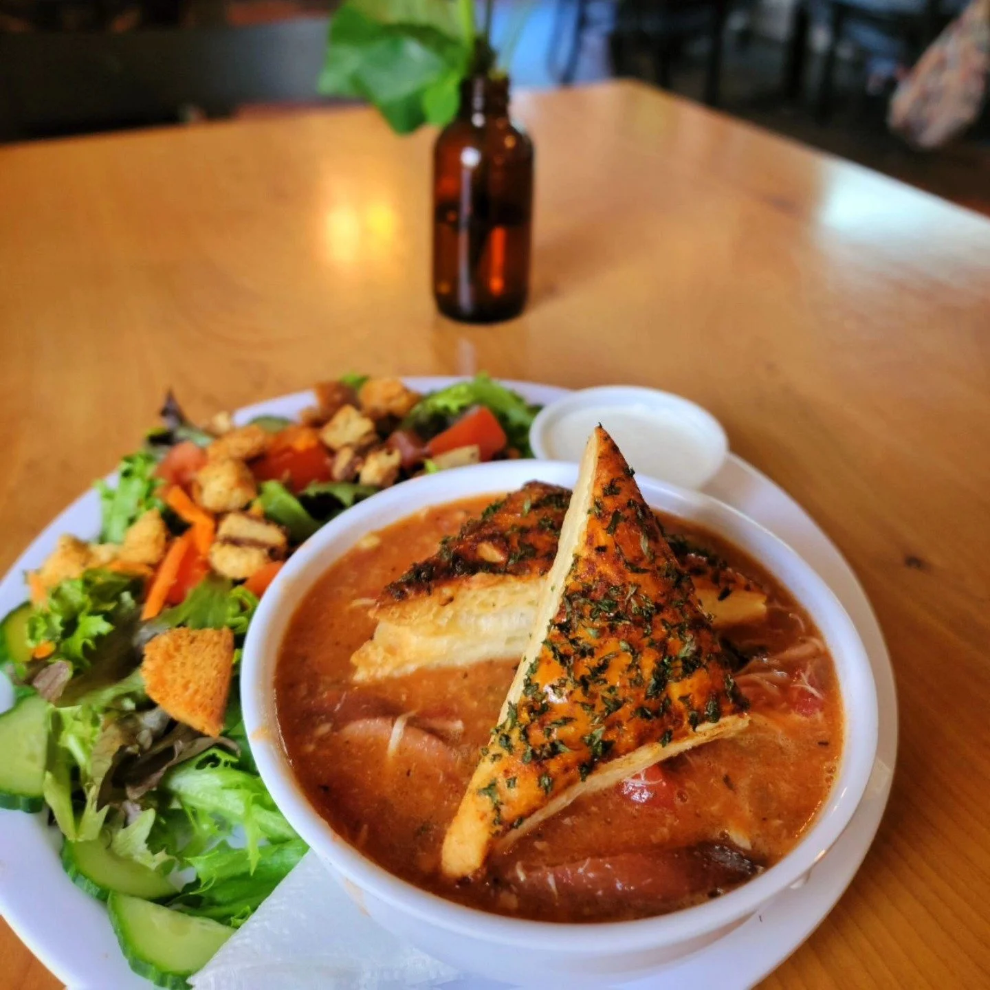 A bowl of tomato-based soup with cheese bread, a side salad with croutons, and a small container of sour cream on a wooden table.