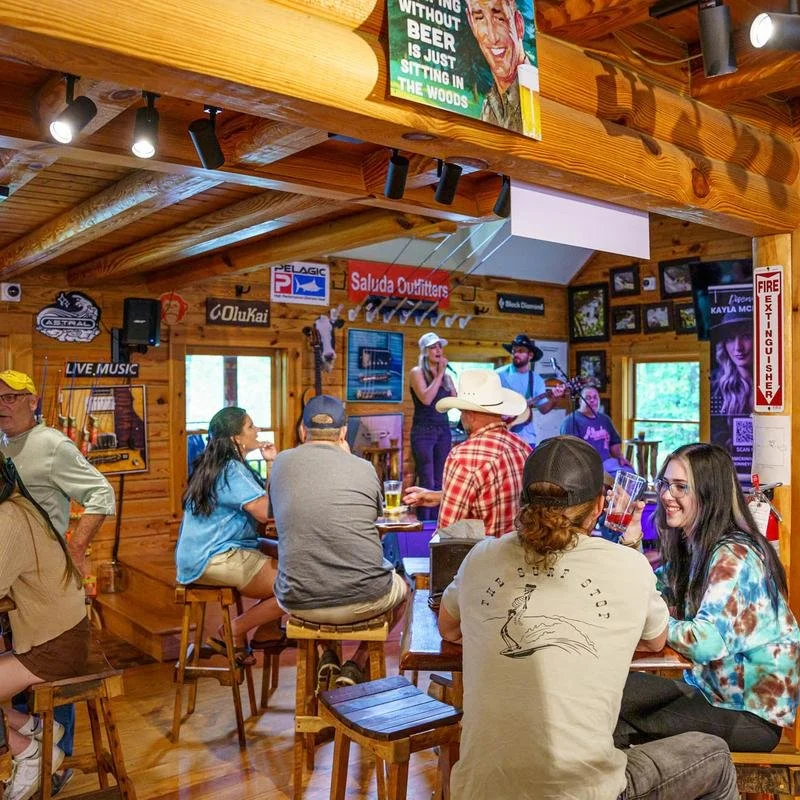 People sitting at a wooden bar watching a live band perform on stage inside a cozy, wood-paneled pub or restaurant, with many signs and framed photos on the walls and a woman singing into a microphone.