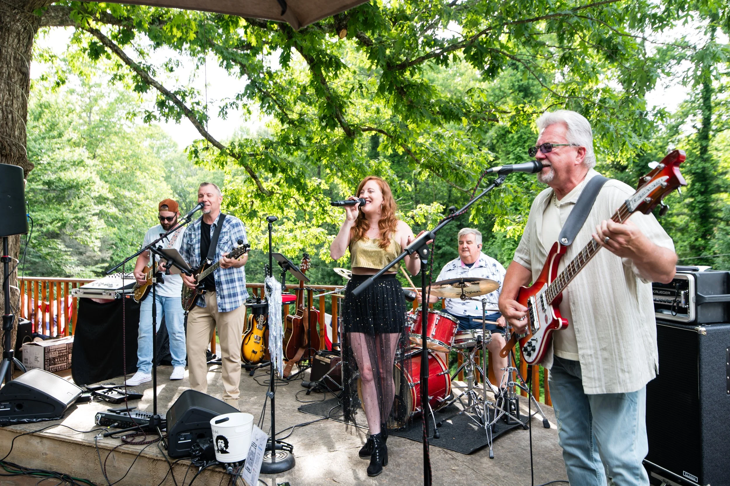 A group of musicians performing outdoors on a stage under trees, including a female singer, guitarists, and a drummer.