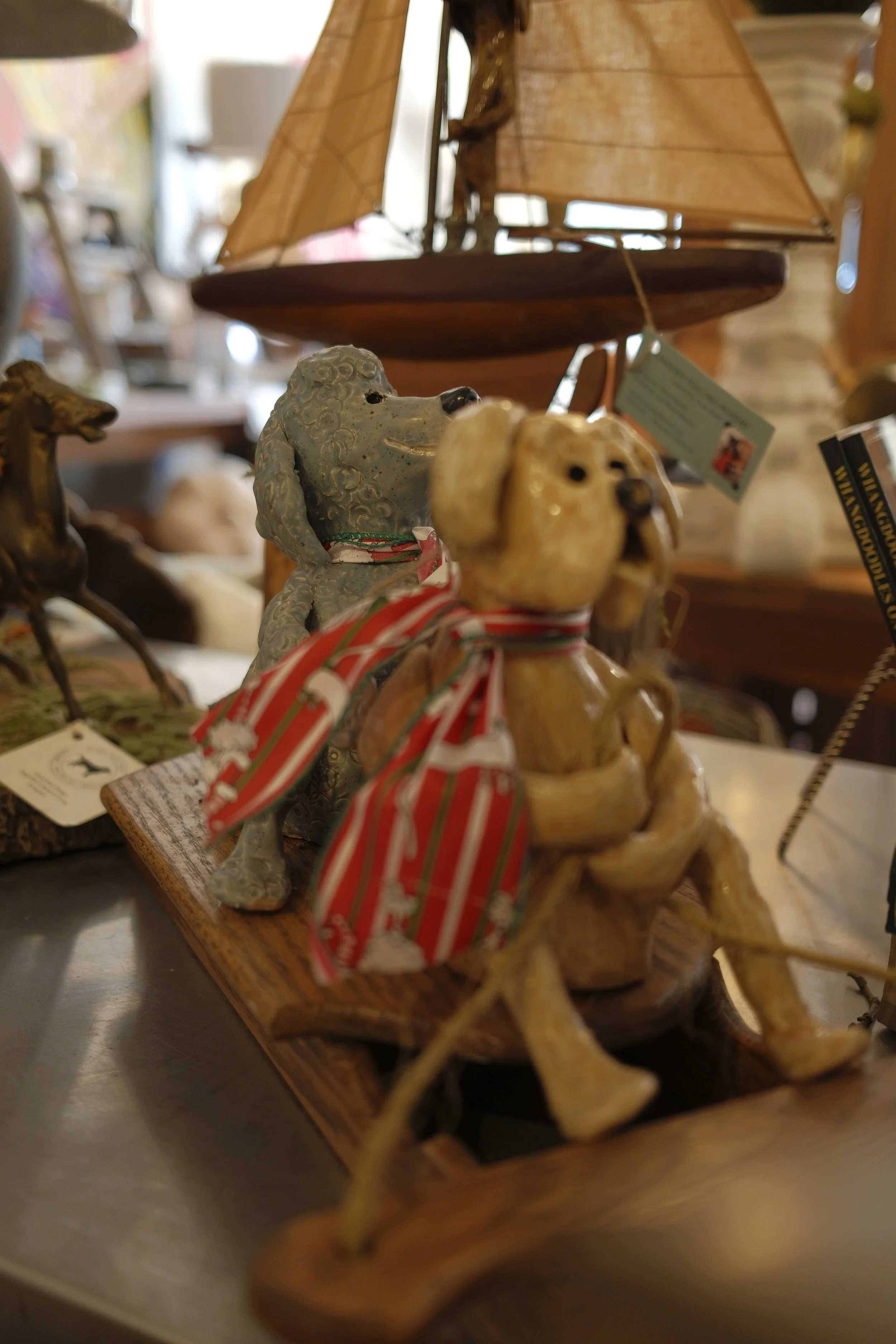 Wooden sailboat model with tan sails and navy blue and gold accents, and ceramic animal figurines, including a yellow dog with a red and green striped ribbon and a gray dog with a white ribbon with red details, displayed on a wooden base.