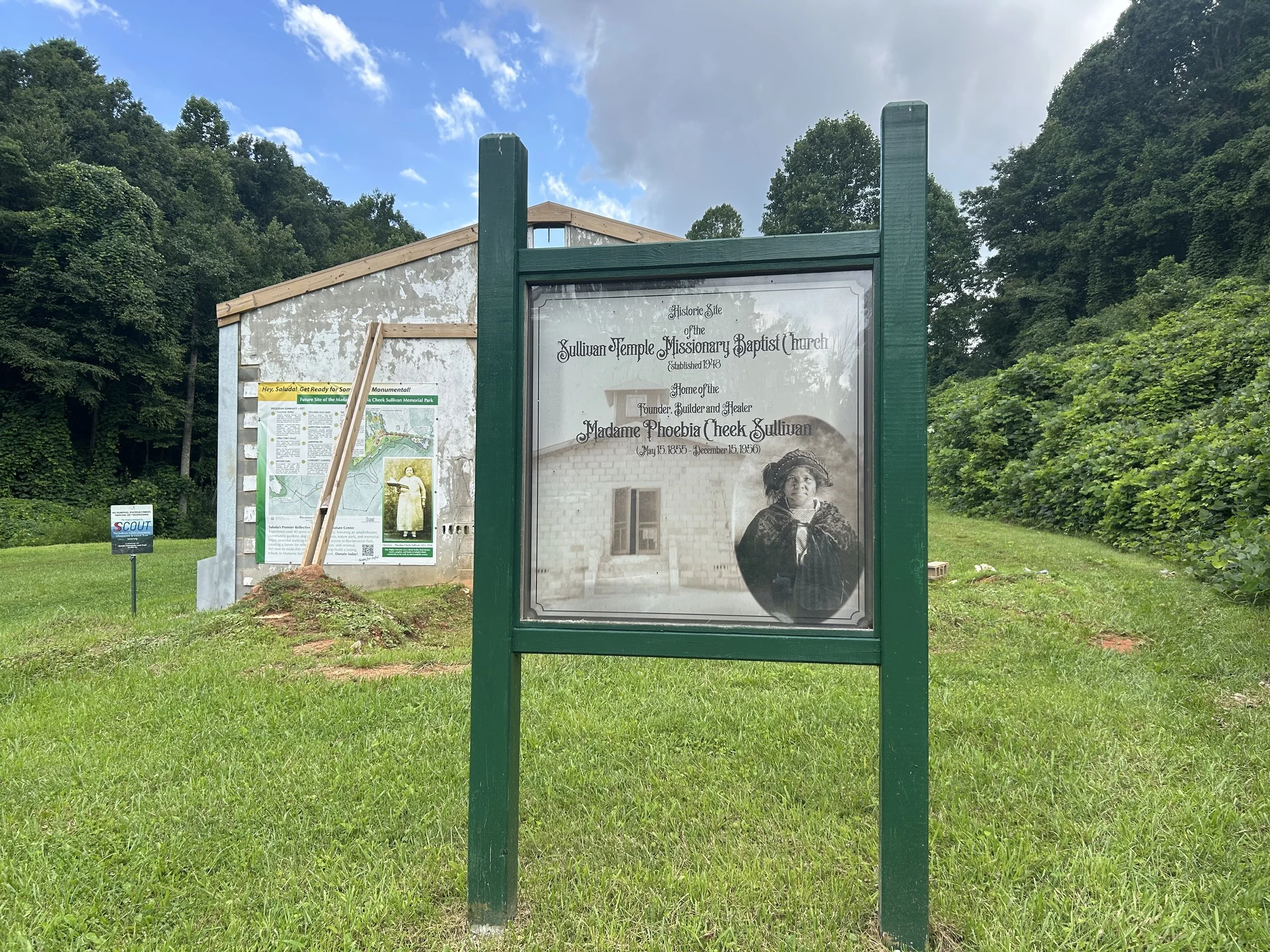 Historical marker in a park with informational plaques and signs, surrounded by green trees and grass under a partly cloudy sky.