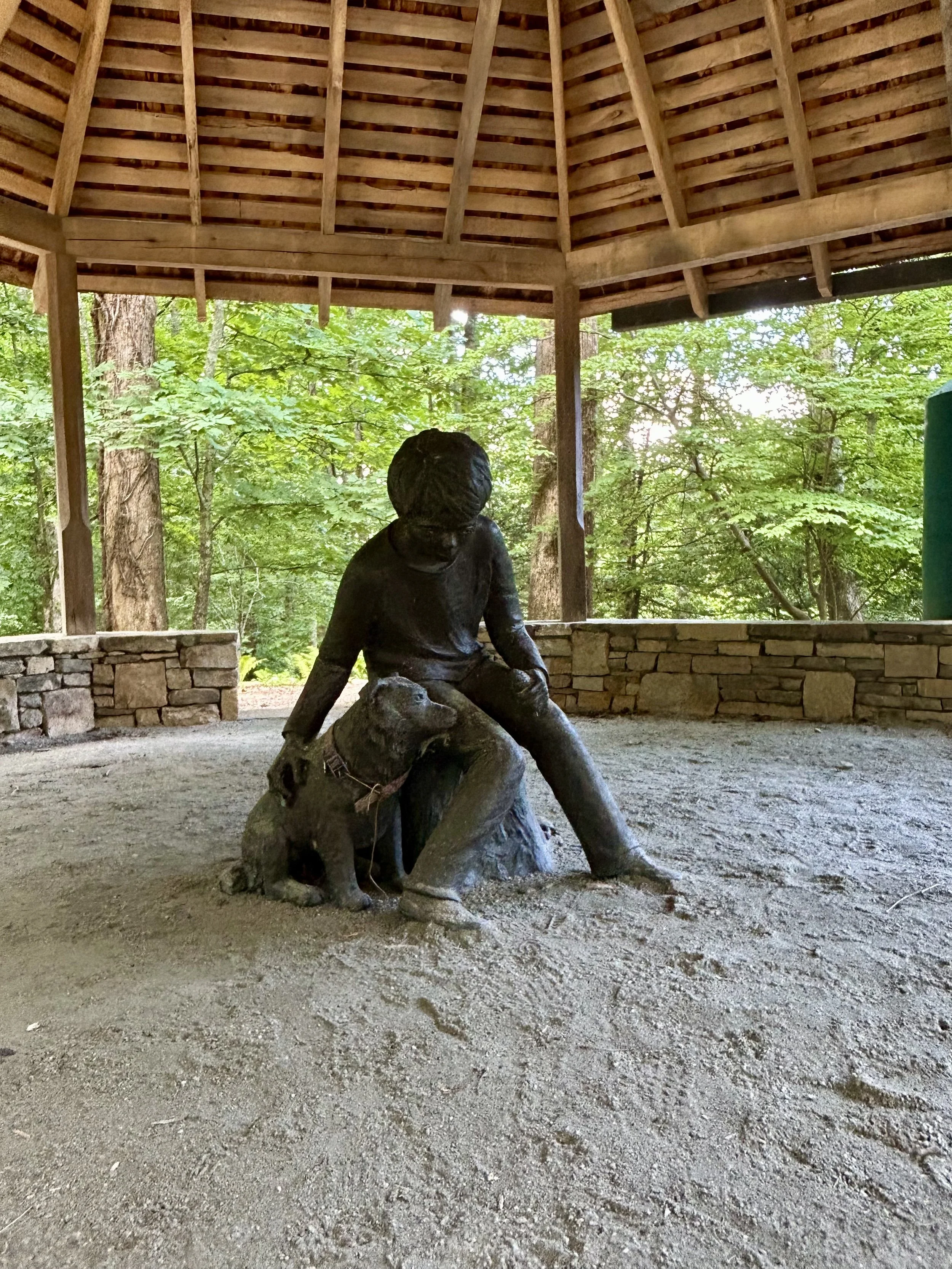 A sculpture of a person riding a pig on a dirt ground inside a wooden open shelter surrounded by green trees.