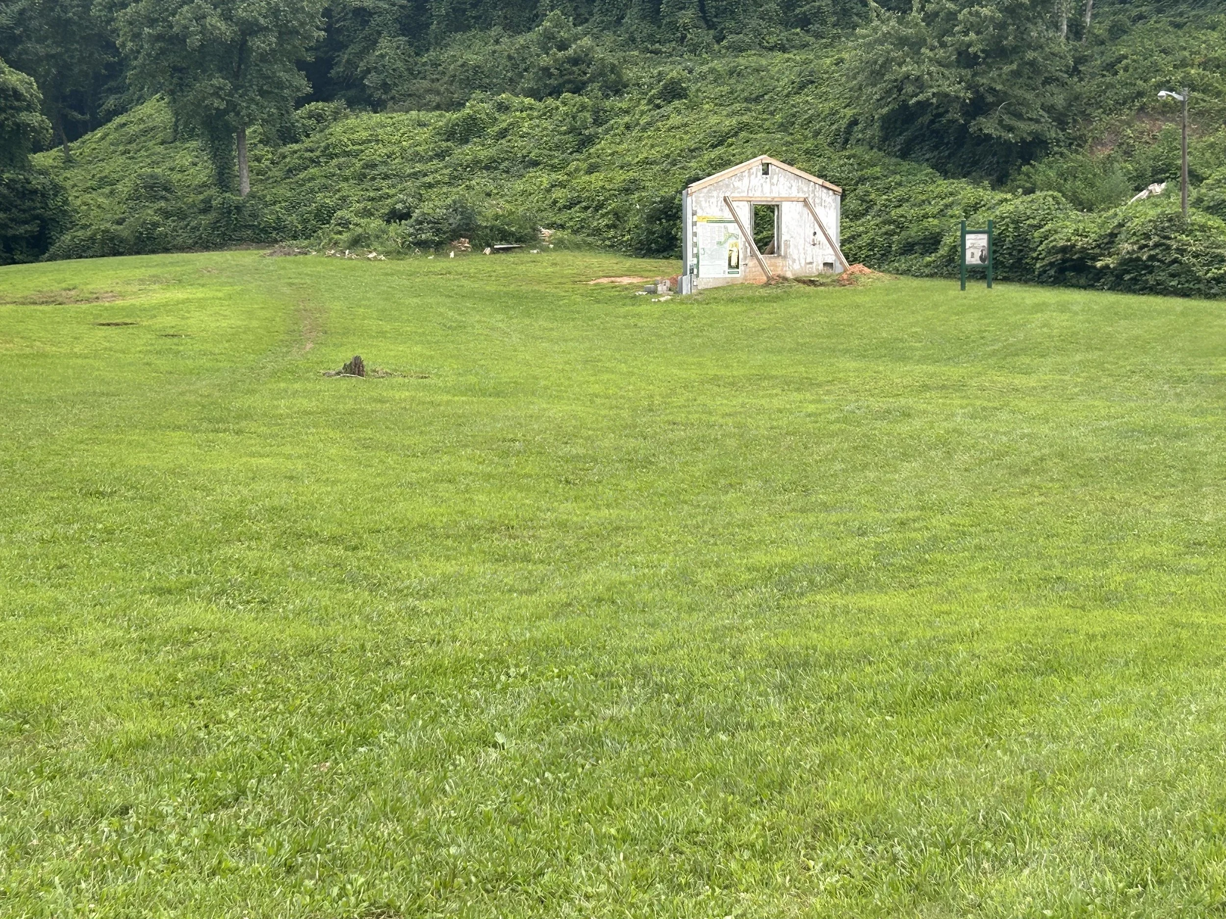 Green grassy field with a small, weathered, white wooden shed and informational signs, surrounded by dense trees and bushes.