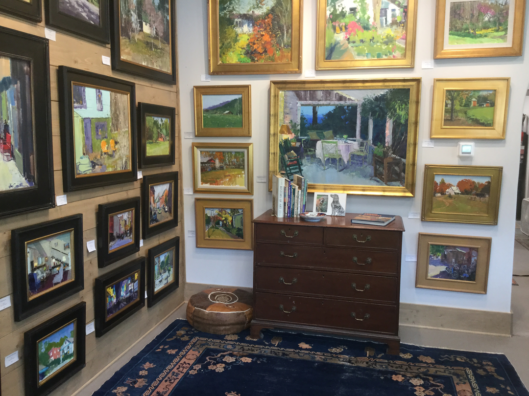 An art gallery corner with multiple landscape paintings in gold and black frames on the walls. A wooden chest of drawers holds books and decorative items. A patterned rug covers part of the floor, and a round pet bed is placed near the chest.