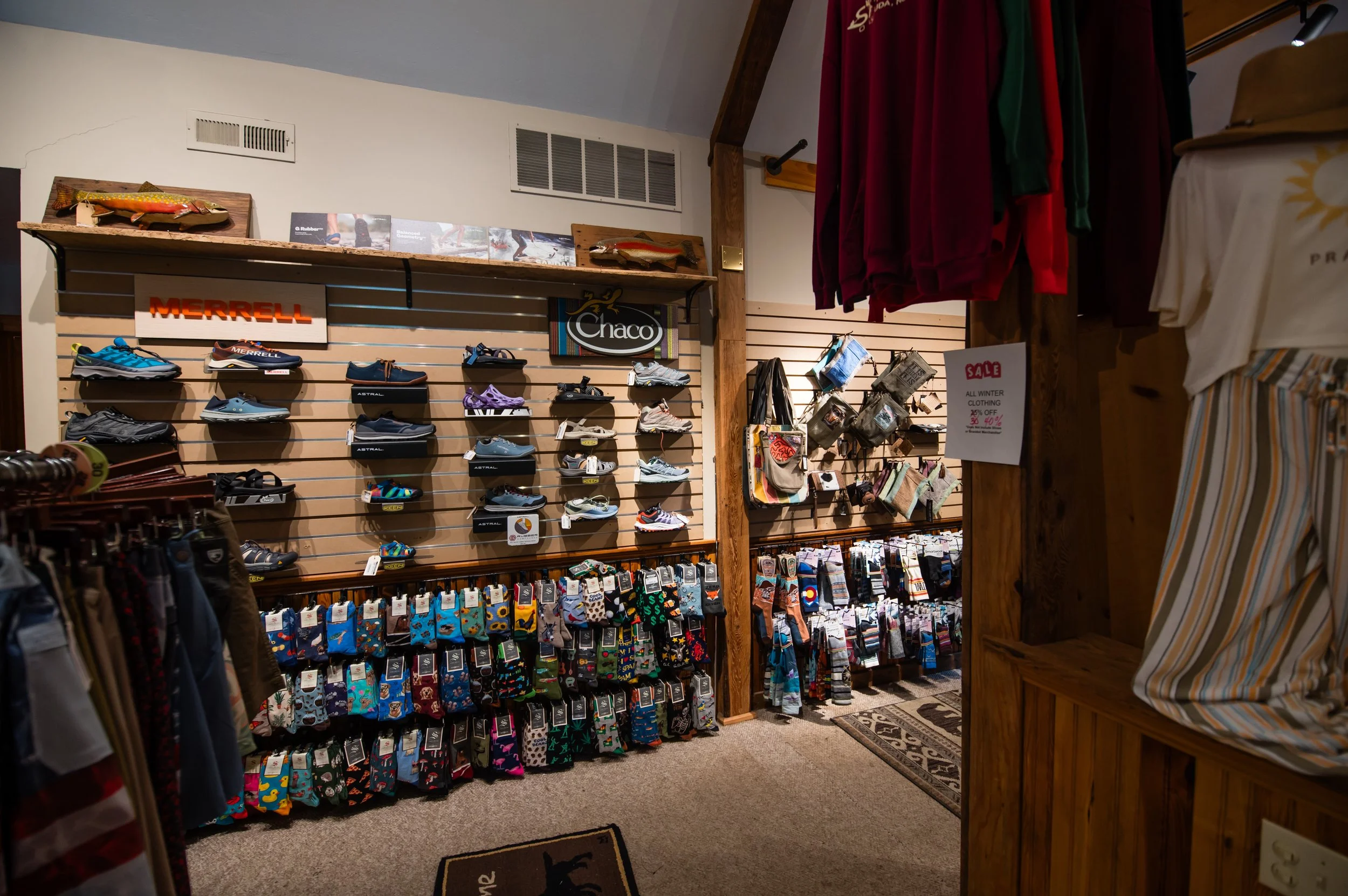 A store section with shoes on display and socks hanging, promotional signs, and clothing on racks, with a wooden and beige interior.