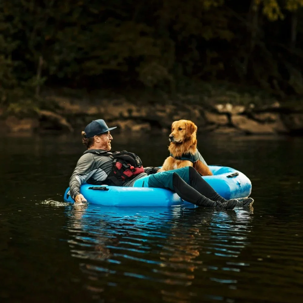 A man and a golden retriever dog sitting on a blue inflatable boat floating on a calm river in a forested area.