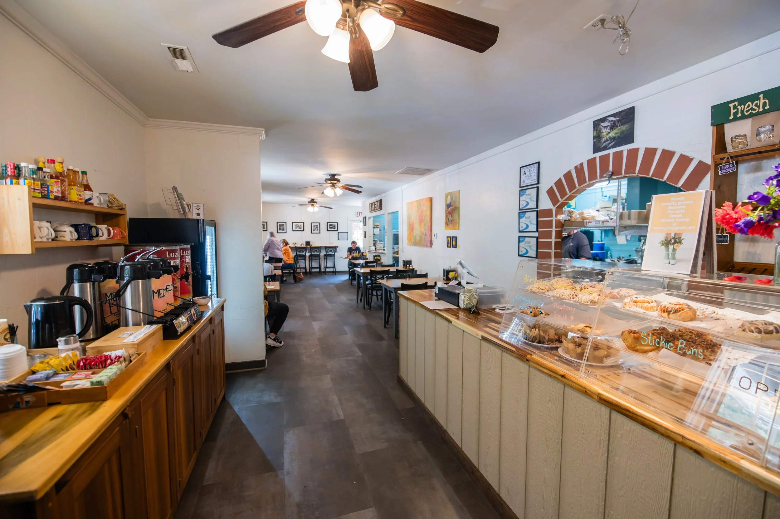 Interior of a small bakery or cafe with a display case filled with cookies and baked goods. The space has wooden accents, a ceiling fan, and artwork on the walls. Customers are seen sitting and dining in the background.
