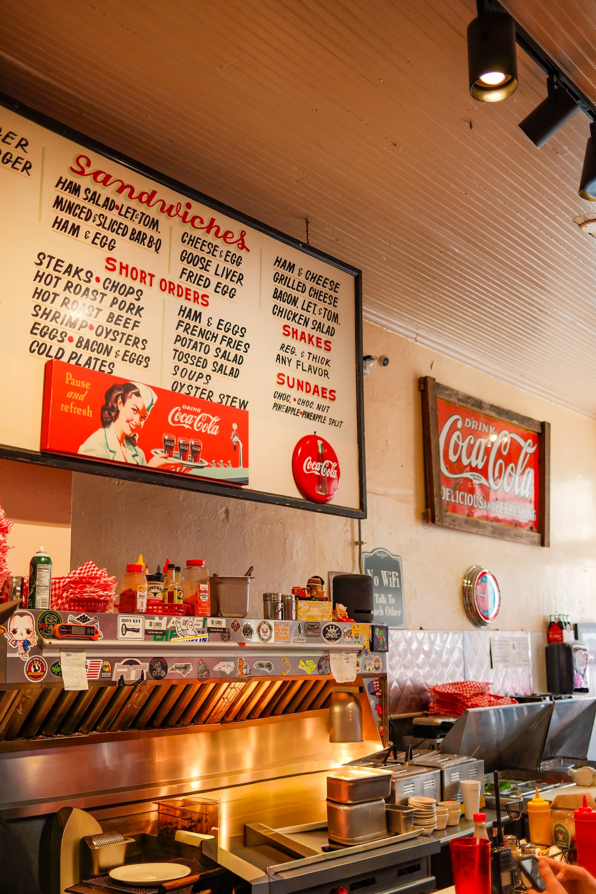 Inside a restaurant kitchen, a menu board hangs on a wall listing sandwiches, short orders, shakes, and sundaes. The counter has various condiments, stickers, and kitchen tools. There is a grill area and a person is holding a phone on the right.