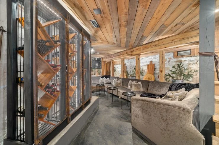 Cozy lounge with wooden ceiling, large windows, plush seating with throw pillows, and a wine storage area with bottles on modern shelves.