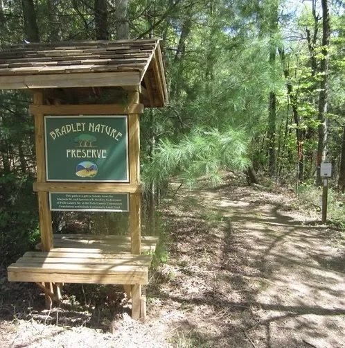 A wooded trail leading into a nature preserve with a sign that reads "Bradley Nature Preserve" and a wooden shelter at the start of the trail.