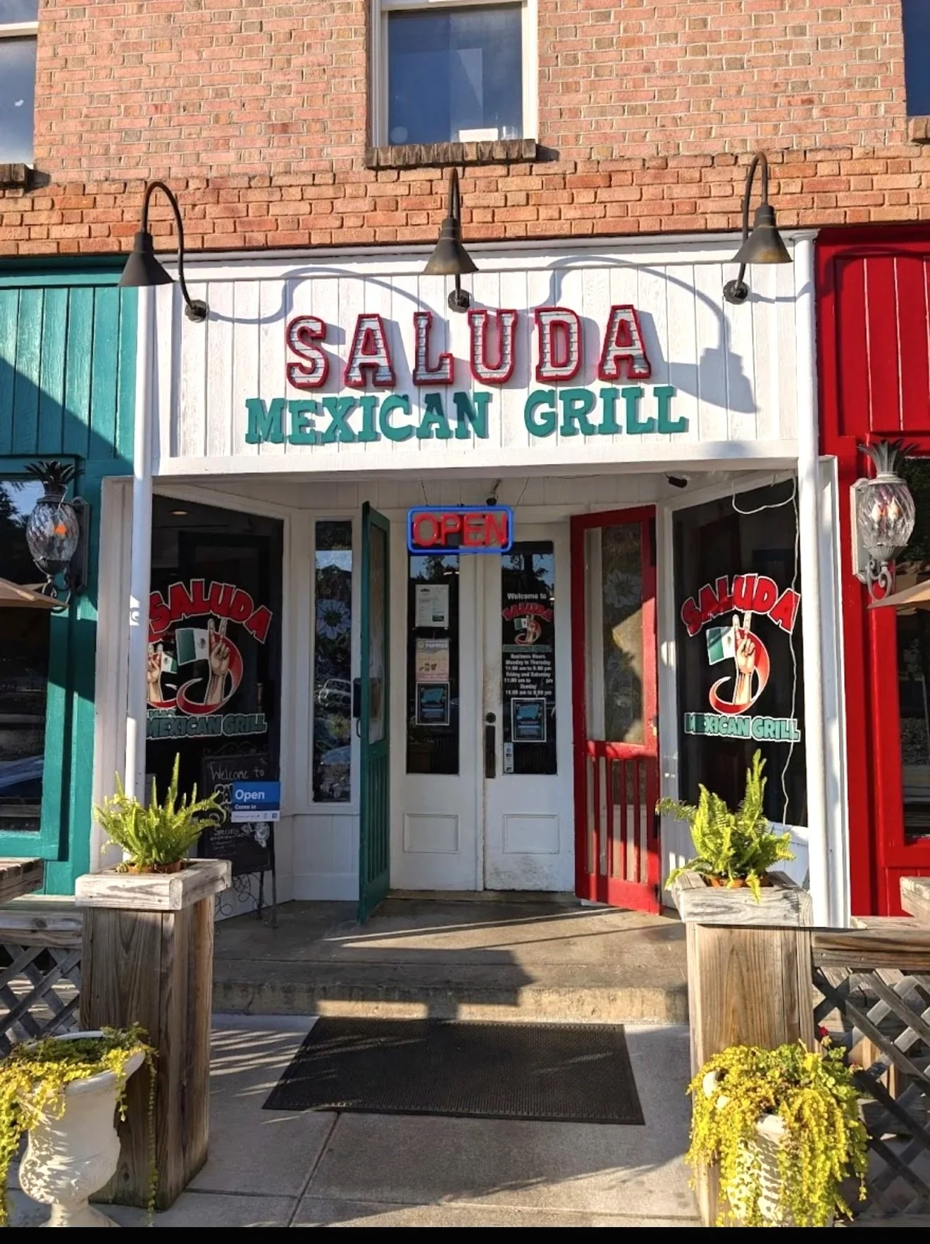 The storefront of Salud Mexican Grill with open sign, two potted plants on wooden planters, and the exterior decorated with rustic red, green, and white colors.