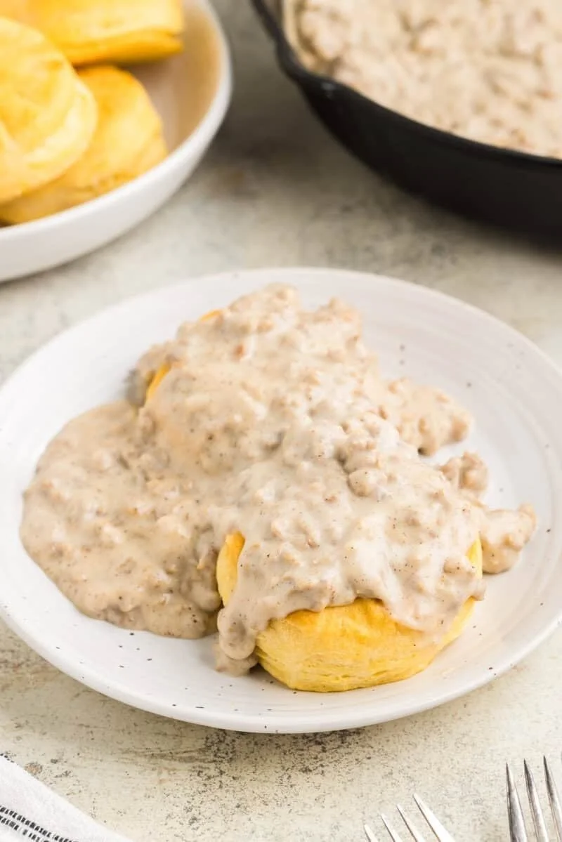 Creamed biscuits with sausage gravy on a white plate, served with potato chips and another dish in the background.
