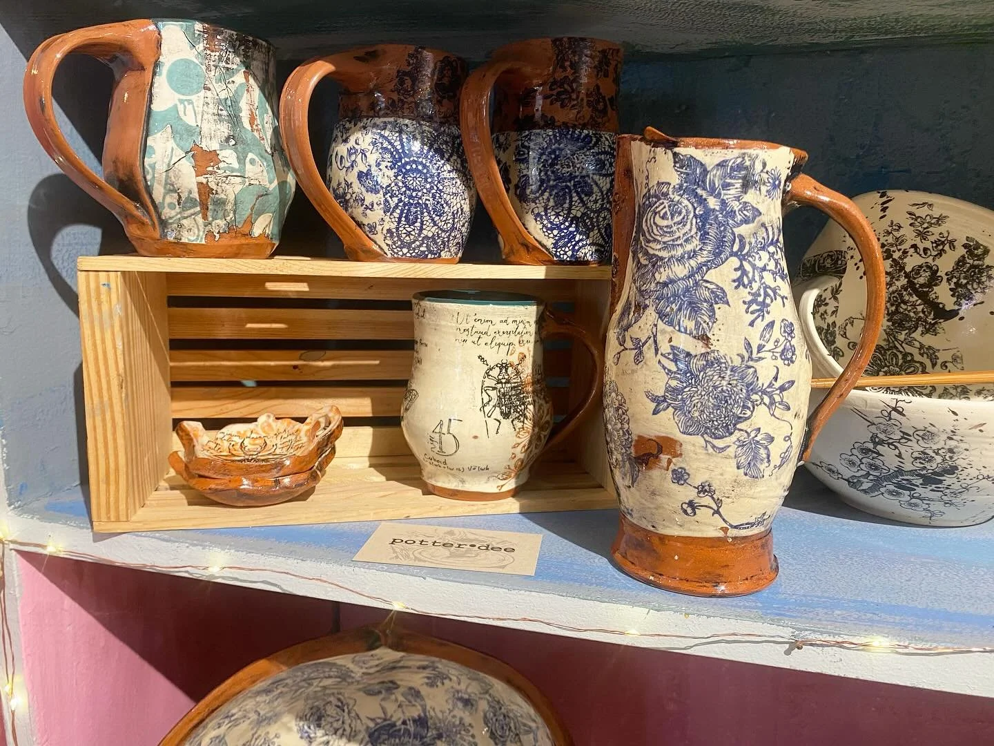 Collection of ceramic mugs and a small bowl with blue and beige floral design, displayed on a wooden shelf and crate.