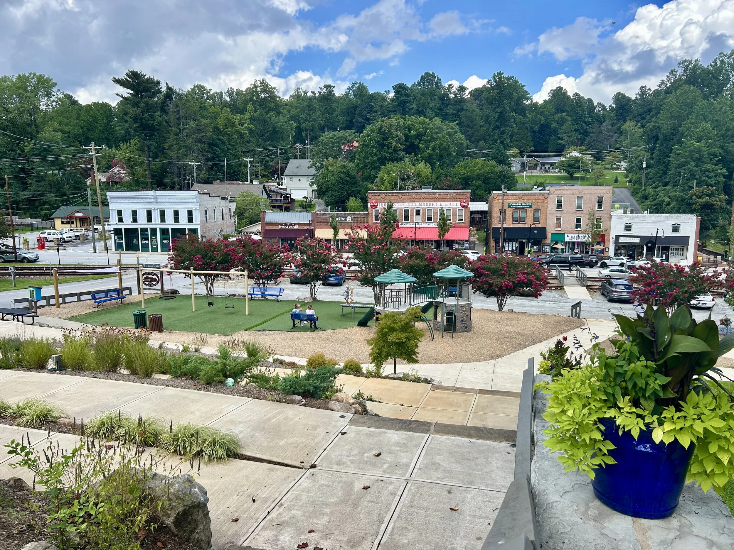 View of small town with colorful storefronts, blooming pink trees, parking lot, and hillside with houses and trees under a blue sky with clouds.