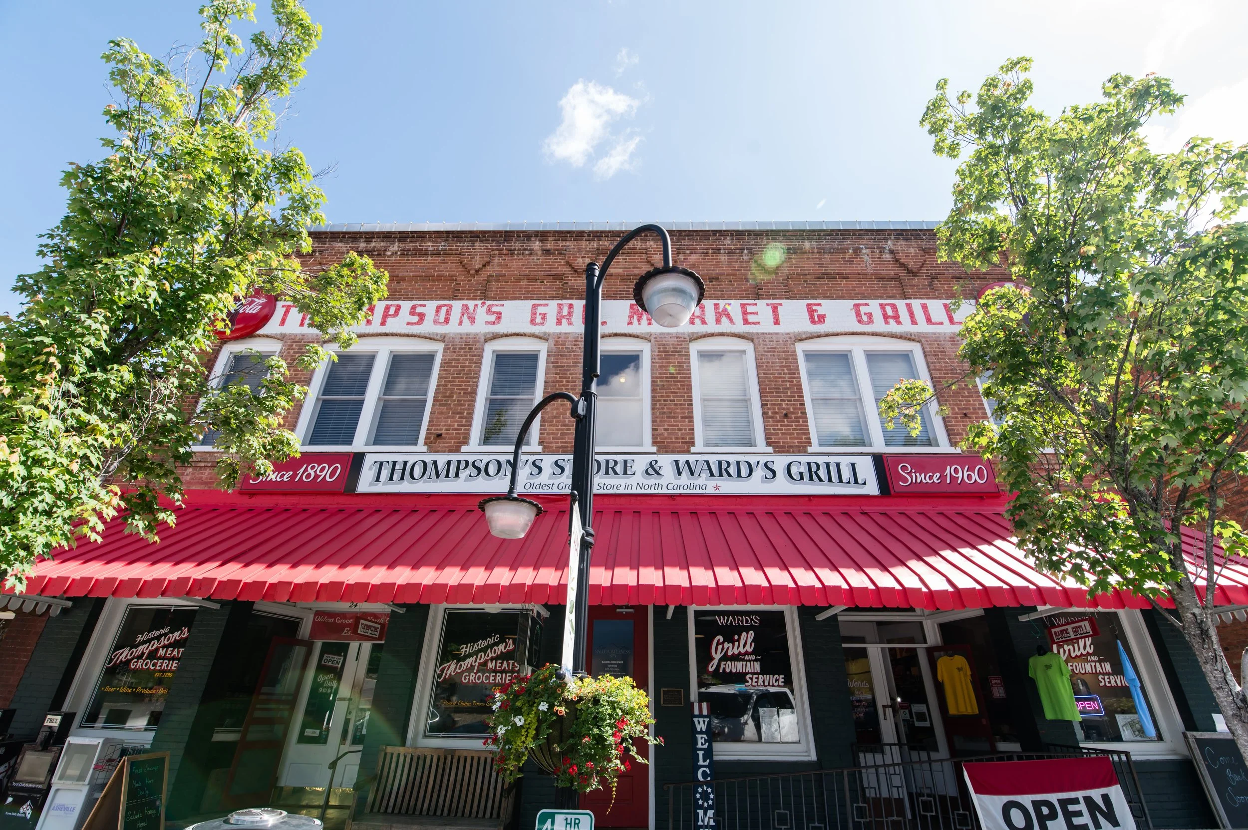 The image shows a brick building with a red and white awning. The sign on the building reads 'Thompson's Store & Ward's Grill.' There are several windows above the storefront, and a street lamp is in the center foreground. Two trees flank the buildin