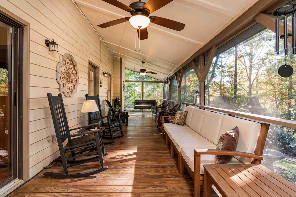 A covered porch with wooden flooring, beige siding, wooden rocking chairs, a white cushioned bench, and a ceiling fan.