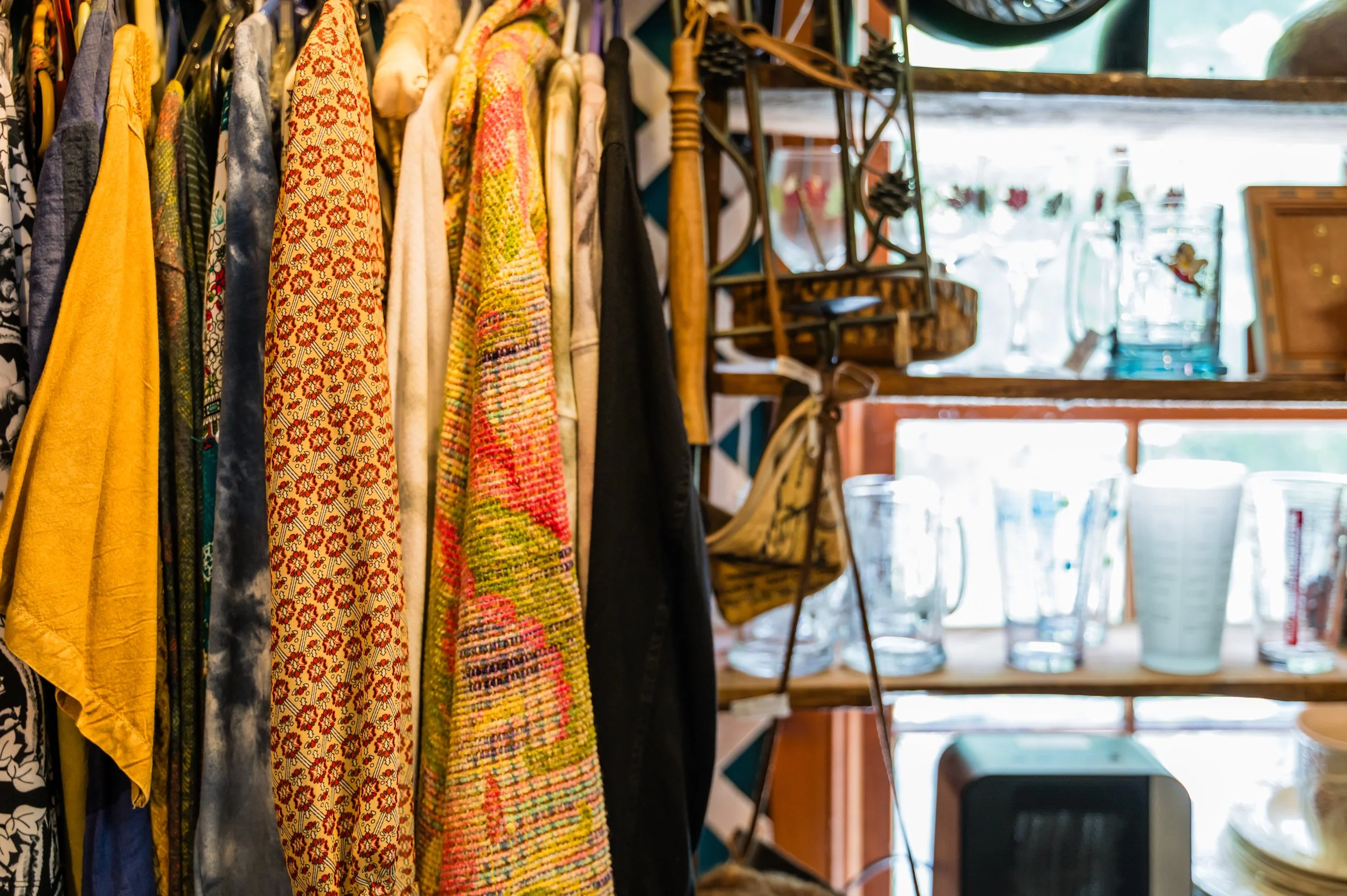 Colorful scarves hanging on a rack in a store, with shelves and windows in the background.