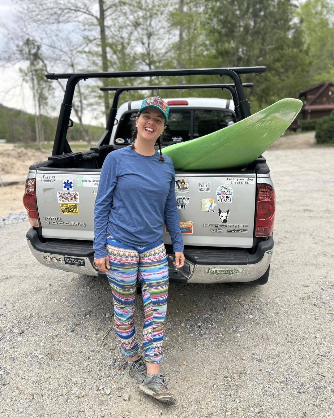 A woman standing in front of a pickup truck with a green surfboard in its bed, smiling at the camera. The woman is wearing a blue long-sleeve shirt, colorful striped pants, and a colorful cap. The truck is decorated with various stickers.