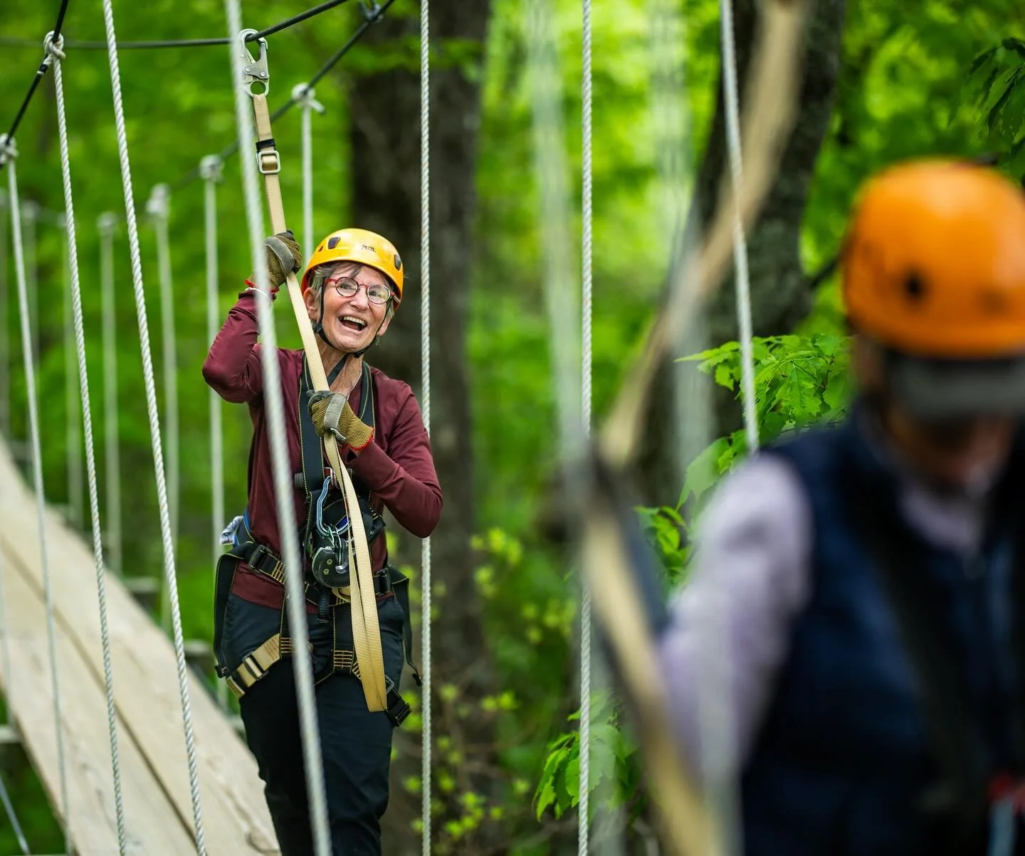An elderly woman smiling and laughing on a suspension bridge in a forest, wearing a yellow helmet and safety harness, with another person partially visible in the foreground.